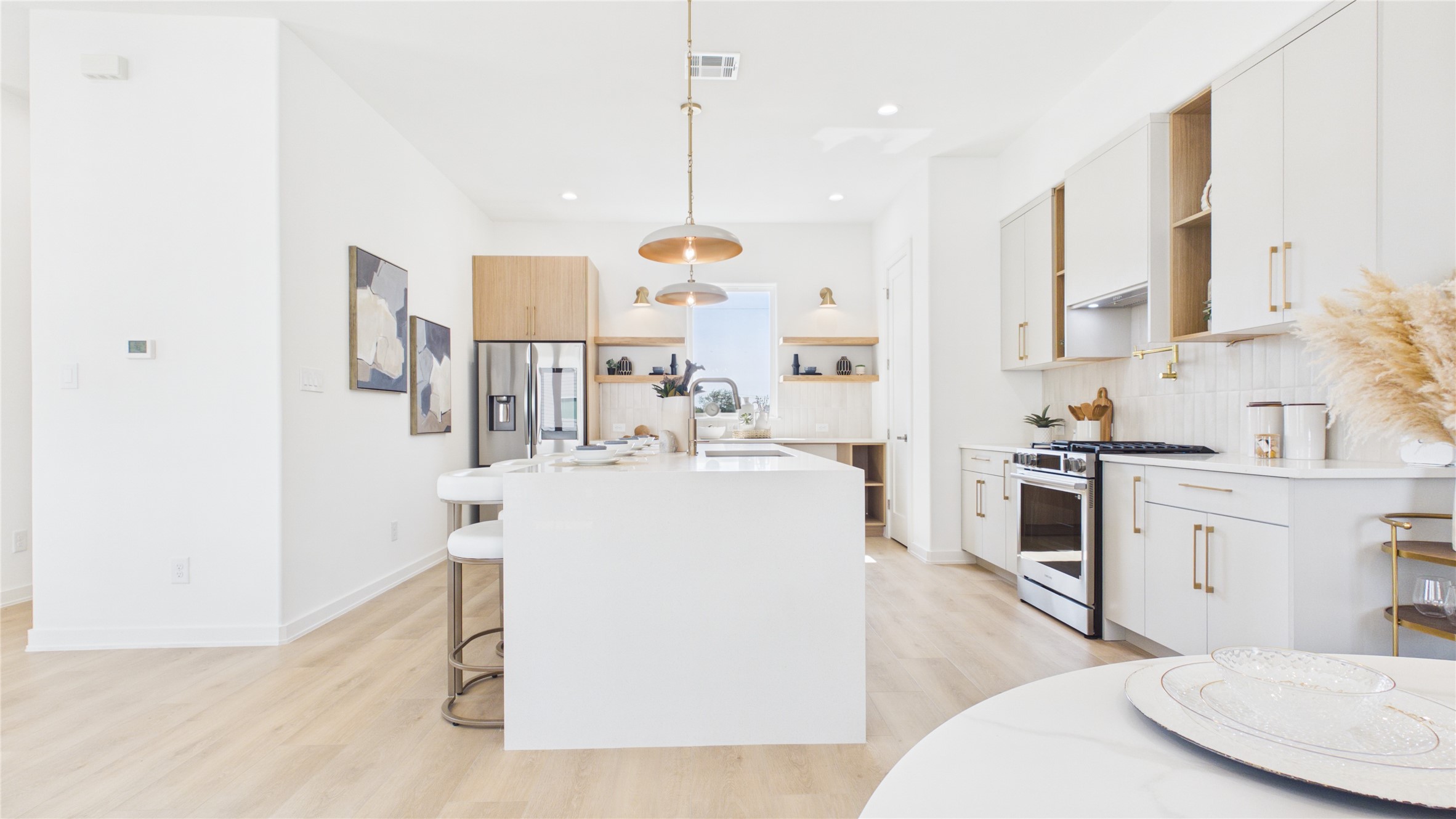 Bright and modern kitchen featuring a sleek island with bar seating, stainless steel appliances, and elegant pendant lighting. The open layout and light wood flooring enhance the spacious feel.
