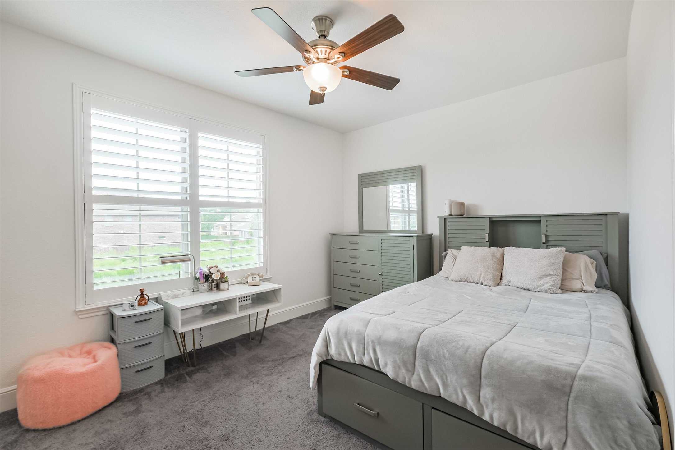 Bright and inviting sixth bedroom featuring a large window with elegant plantation shutters, a ceiling fan, and a warm neutral palette with an en suite bathroom. Abundant natural light creates a serene and comfortable retreat ideal for relaxation.