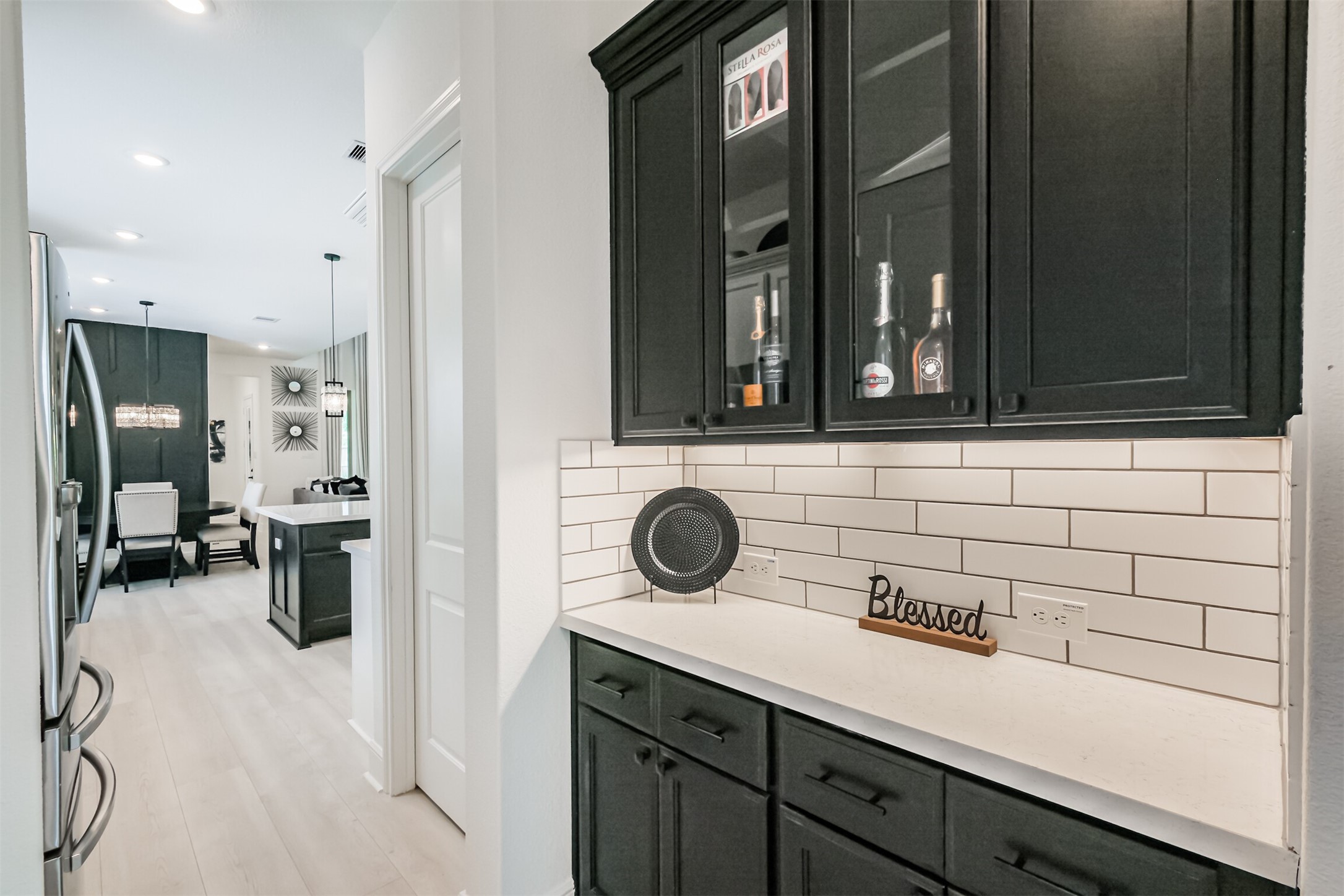Sophisticated butlers pantry featuring sleek black cabinetry with elegant glass-front accents, a crisp white subway tile backsplash, and undercabinet lightingcreating a polished and functional space ideal for entertaining and additional storage.