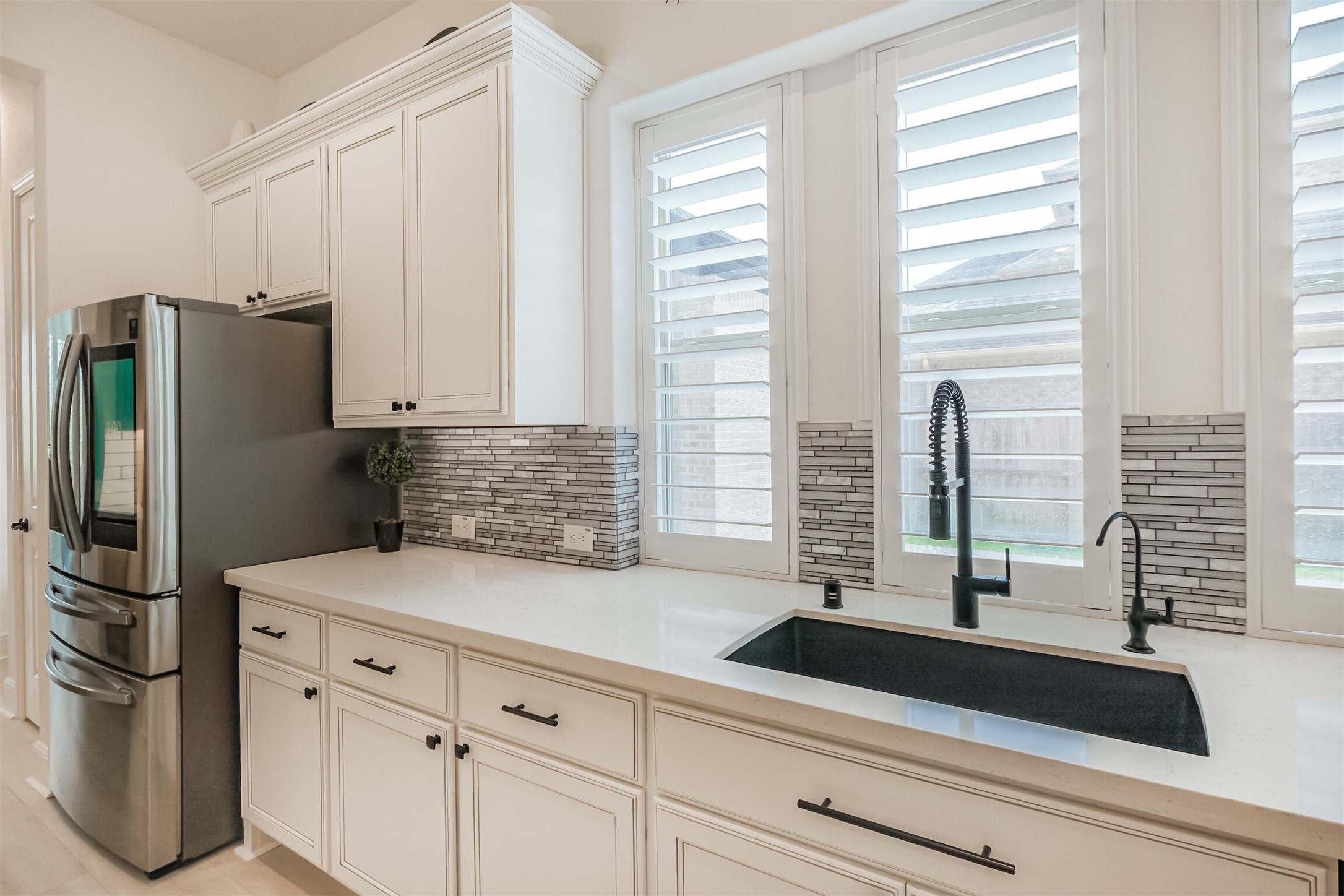 Sophisticated kitchen sink area featuring a sleek undermount black sink with a touchless modern faucet, crisp white cabinetry, and stainless steel appliances. Abundant natural light pours through large windows accented by plantation shutters, while a reverse osmosis and water filtration system adds everyday convenience and luxury.