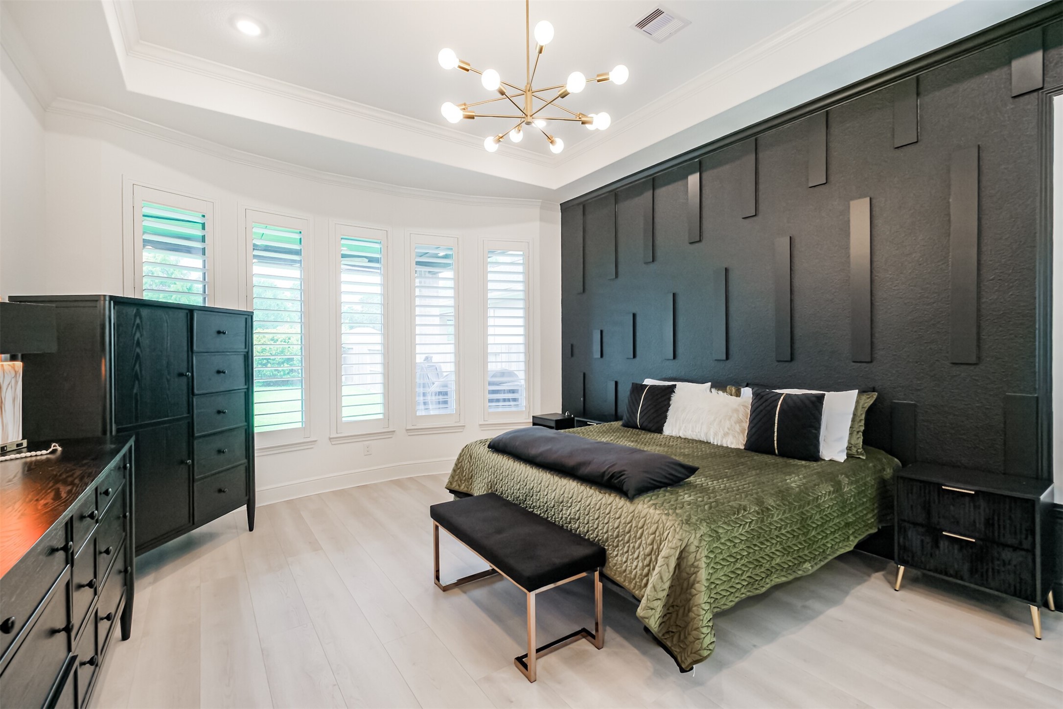 Moving into the luxurious primary suite, a striking custom black accent wall creates a dramatic focal point, complemented by an elegant modern chandelier and tray ceiling. Large windows with plantation shutters fill the space with abundant natural light, creating a sophisticated yet inviting retreat.