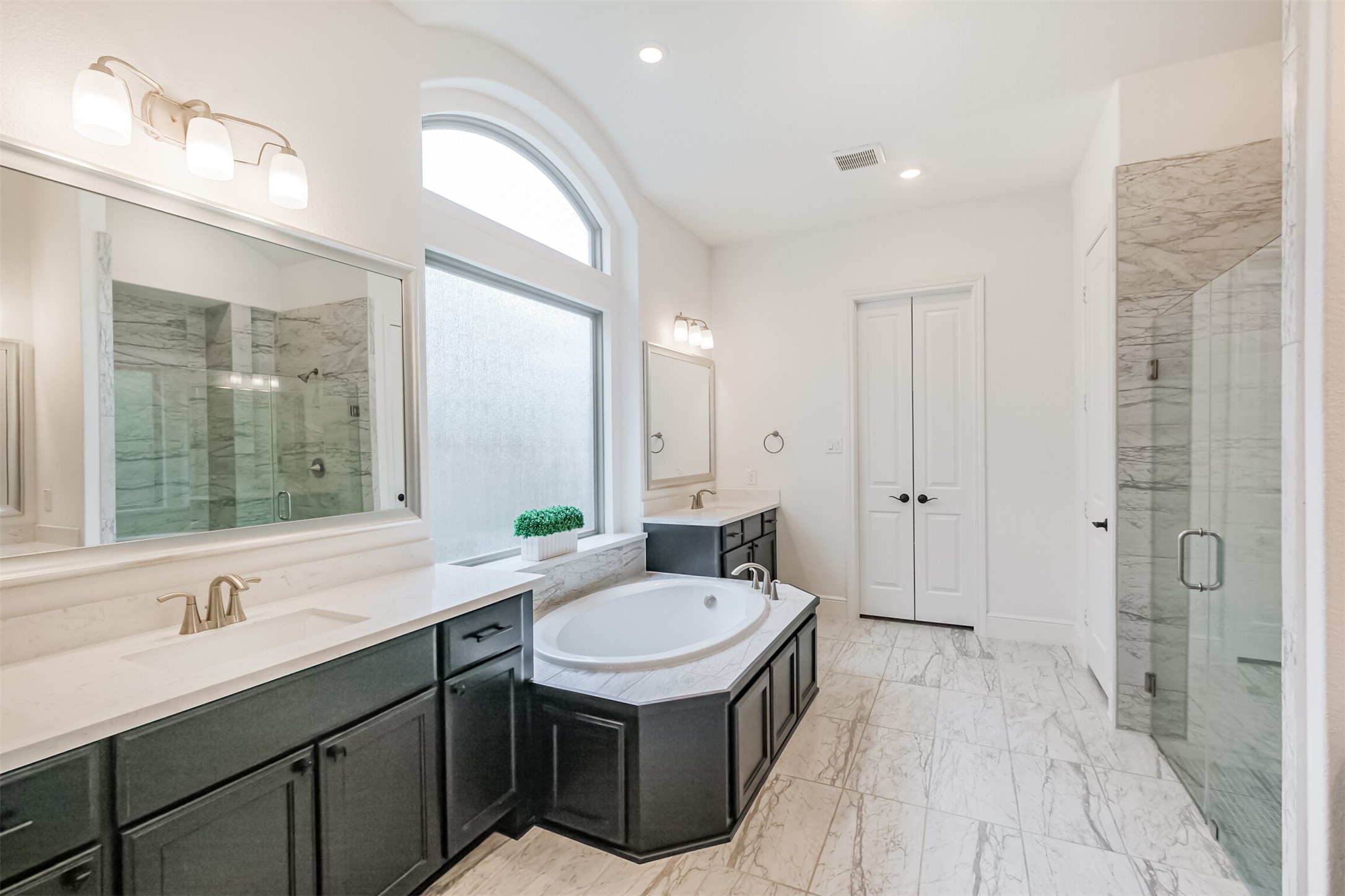 Luxurious primary bathroom designed as a serene retreat, featuring a spacious soaking tub, dual vanities, elegant marble tile flooring, and a glass-enclosed shower. Abundant natural light enhances the bright, spa-like atmosphere and sophisticated finishes throughout.