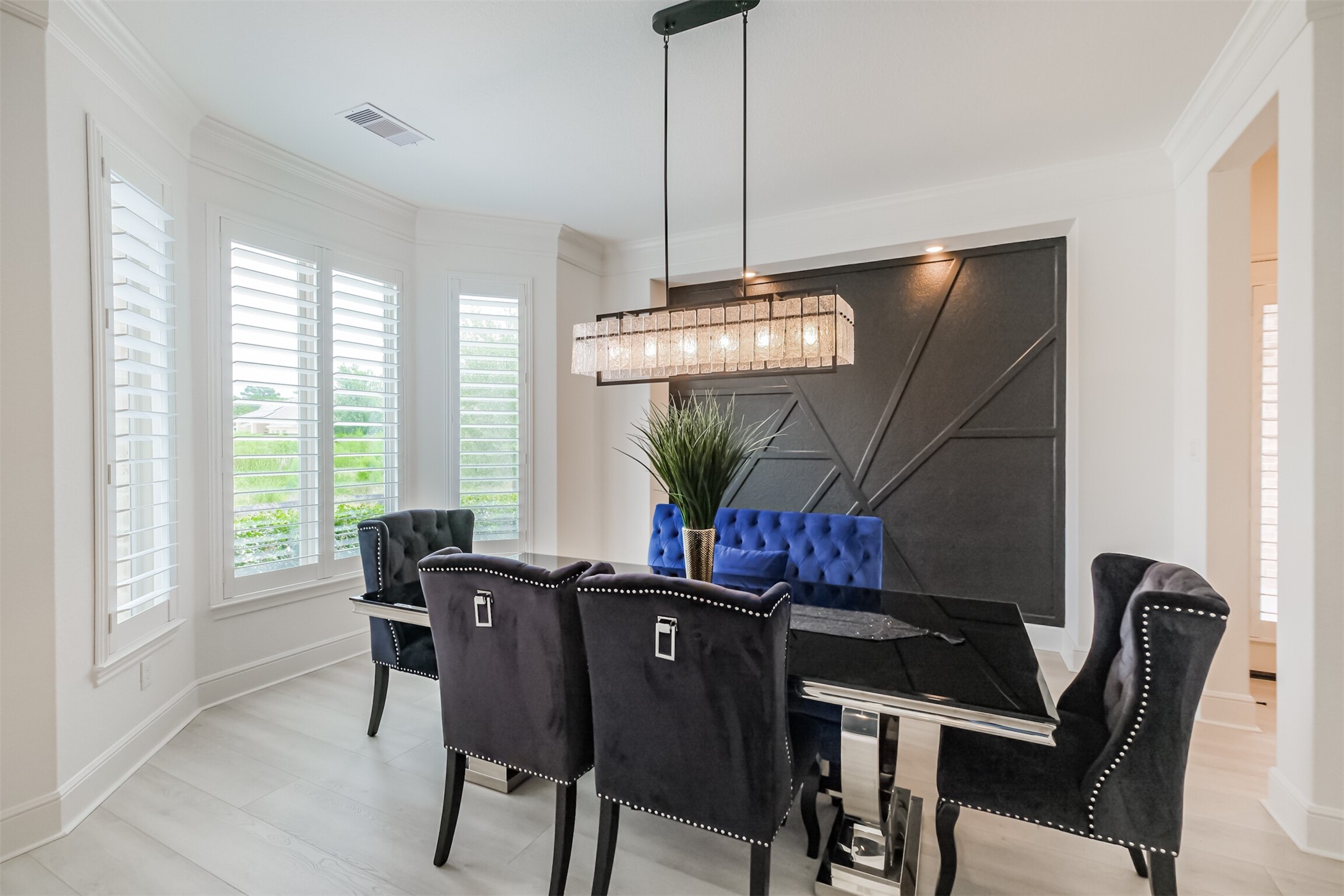Refined formal dining room featuring a striking modern chandelier, custom statement accent wall, and beautiful natural light streaming through expansive windows with plantation shutterscreating a sophisticated setting for elegant dinners and memorable entertaining.