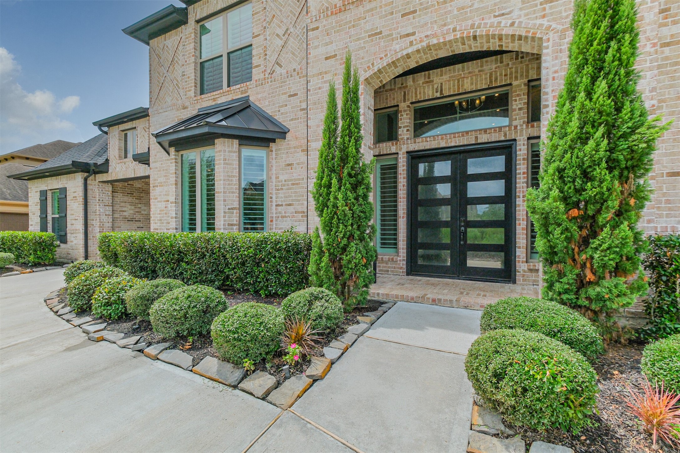 Stunning brick faade featuring elegant double front doors, energy-efficient double-pane windows, and tall, lush greenery complemented by meticulously maintained landscaping for exceptional curb appeal.