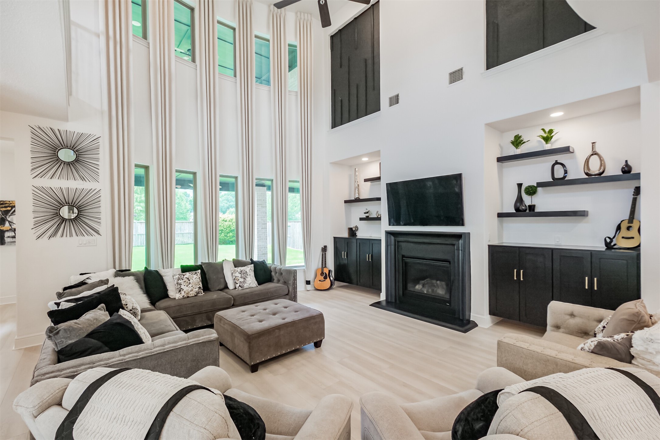 Grand living room showcasing dramatic 25-foot ceilings, expansive bay windows, and abundant natural light. Custom drapery, floating cabinetry and shelving, bespoke architectural accents, and a sleek electric fireplace elevate the space, while impressive 8-foot wood-look plank flooring adds warmth and sophistication throughout.