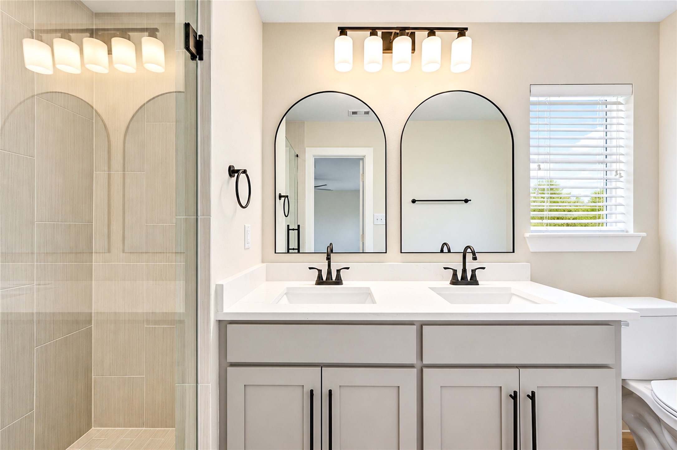 Beautifully remodeled primary bathroom features a dual-sink vanity with elegant Shaker-style cabinetry, soft-close doors, new faucets, arched mirrors, designer lighting, and stylish contemporary finishes.
