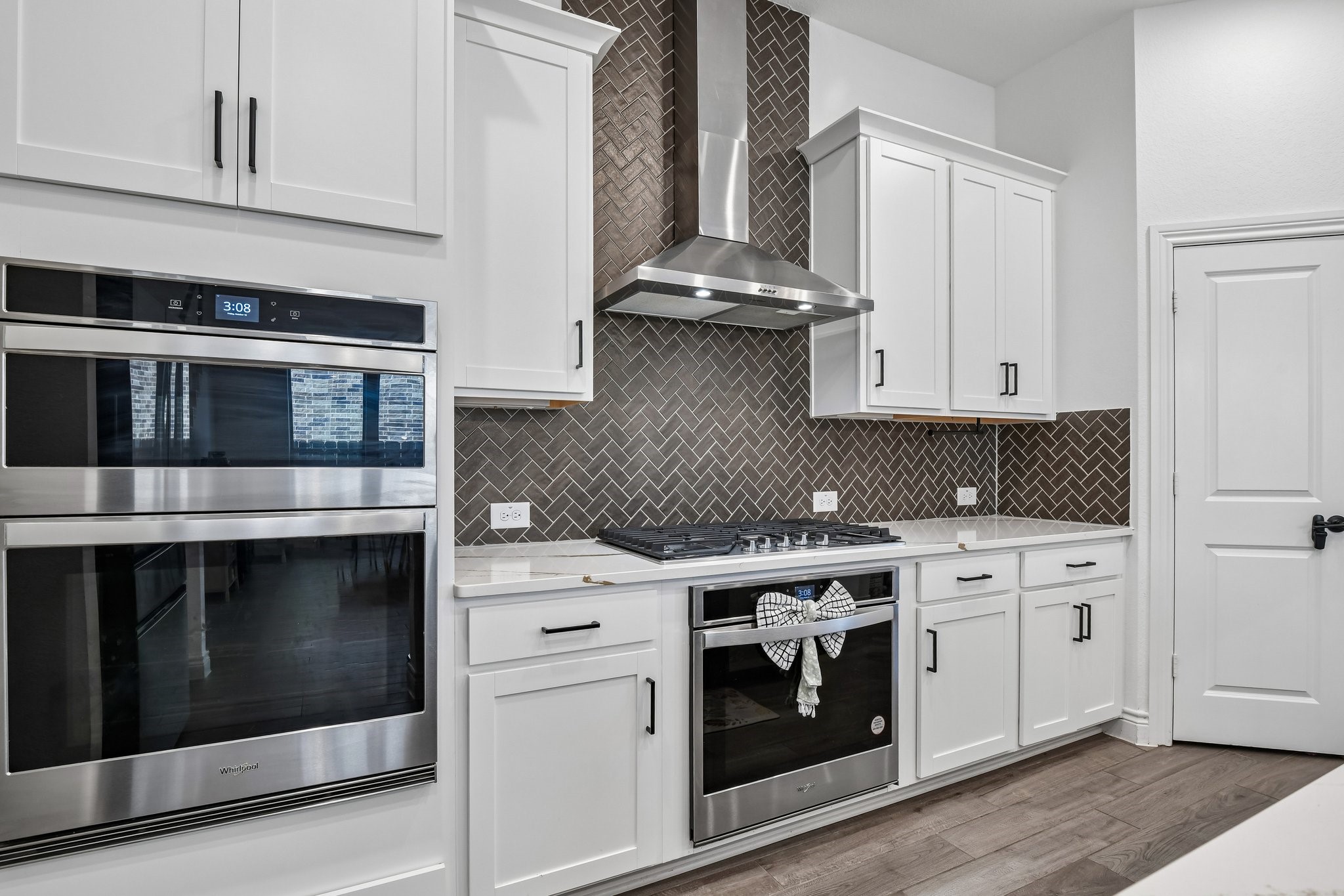 Built-in double ovens, gas cooktop, stainless vent hood, and herringbone tile backsplash