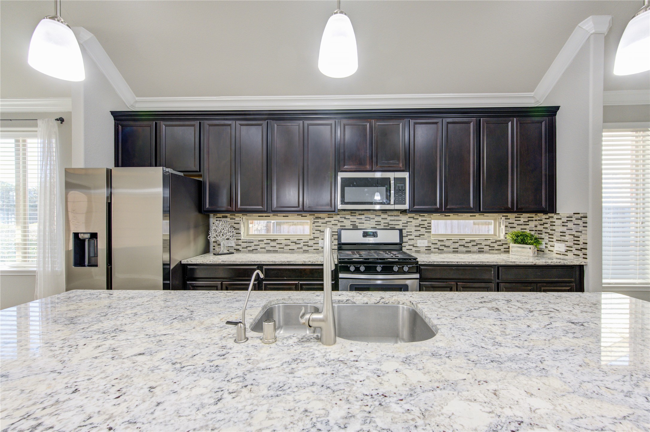 This is a bright, spacious living room with neutral-toned walls and tiled flooring. It features large windows with blinds, a ceiling fan, and a door leading outside. Perfect for a living area or open layout.
