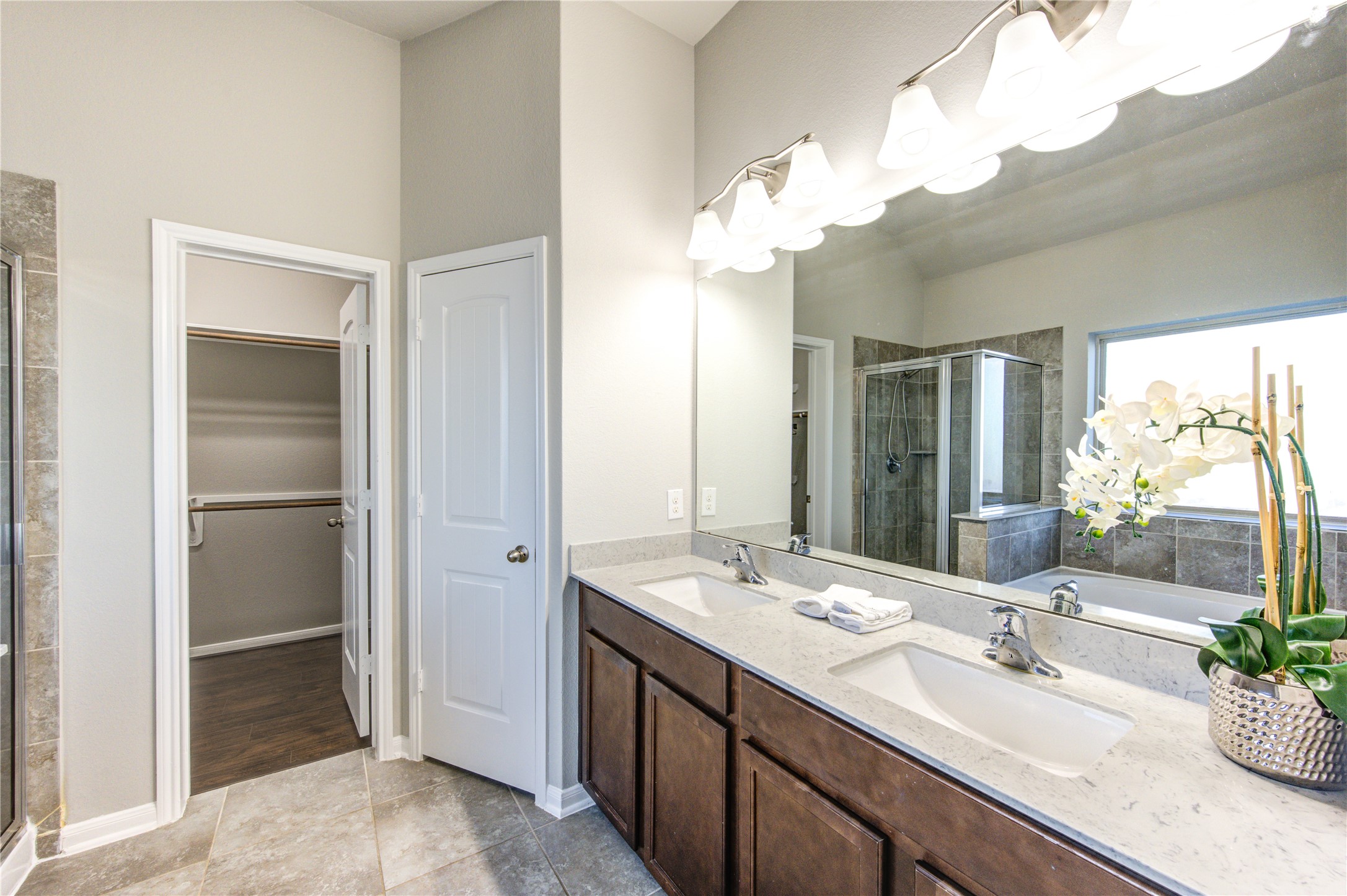This is a modern bathroom with a double vanity featuring dark wood cabinets and a light countertop. It includes a spacious bathtub with tiled surround and large window. The room has a neutral color palette and leads into a room with hardwood flooring.