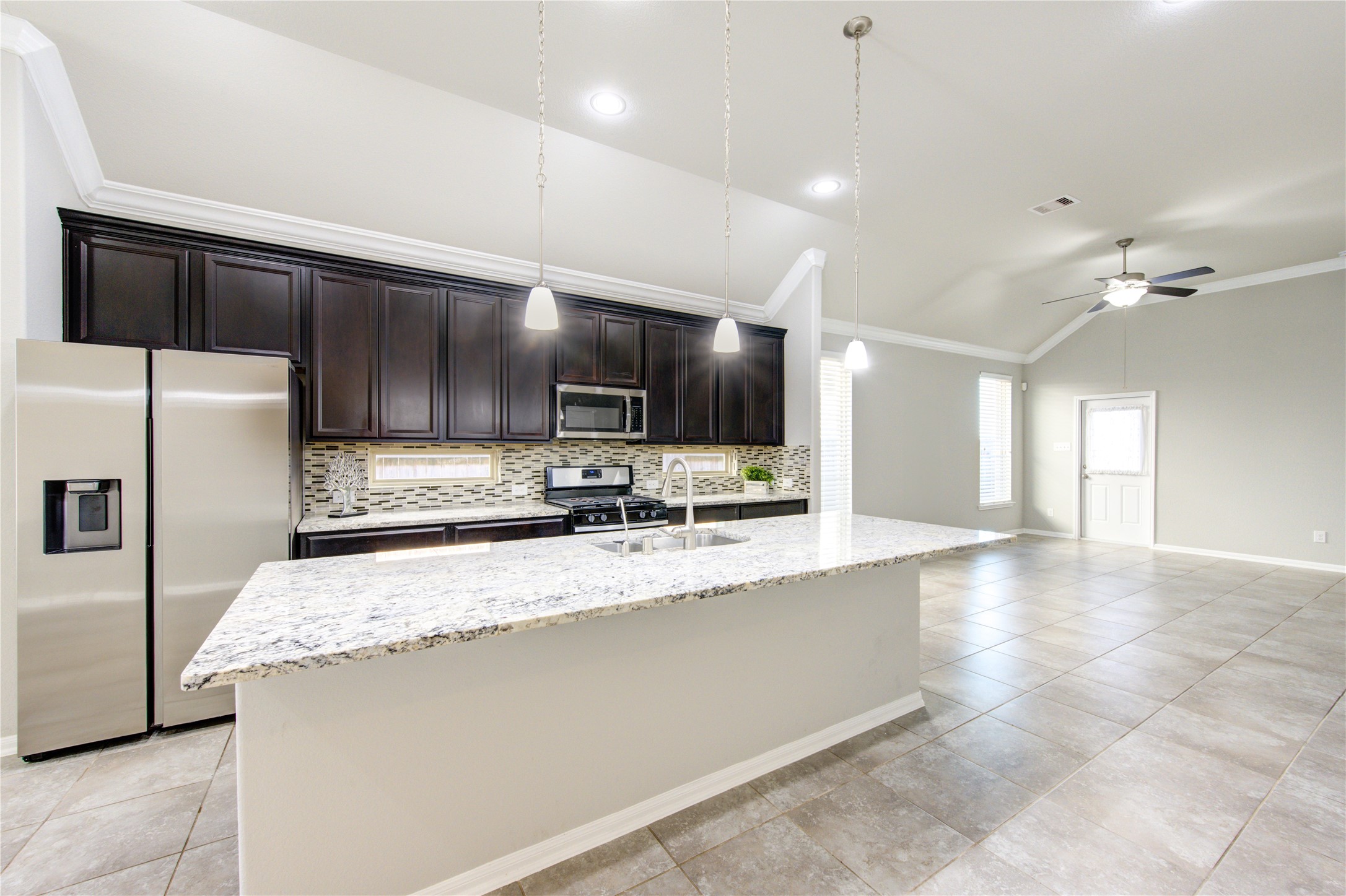 This bright, airy kitchen features large tile flooring, ample natural light from two windows, and a stylish dark wood front door with decorative glass. The space includes a modern stainless steel refrigerator and is accented by crown molding and neutral wall colors.