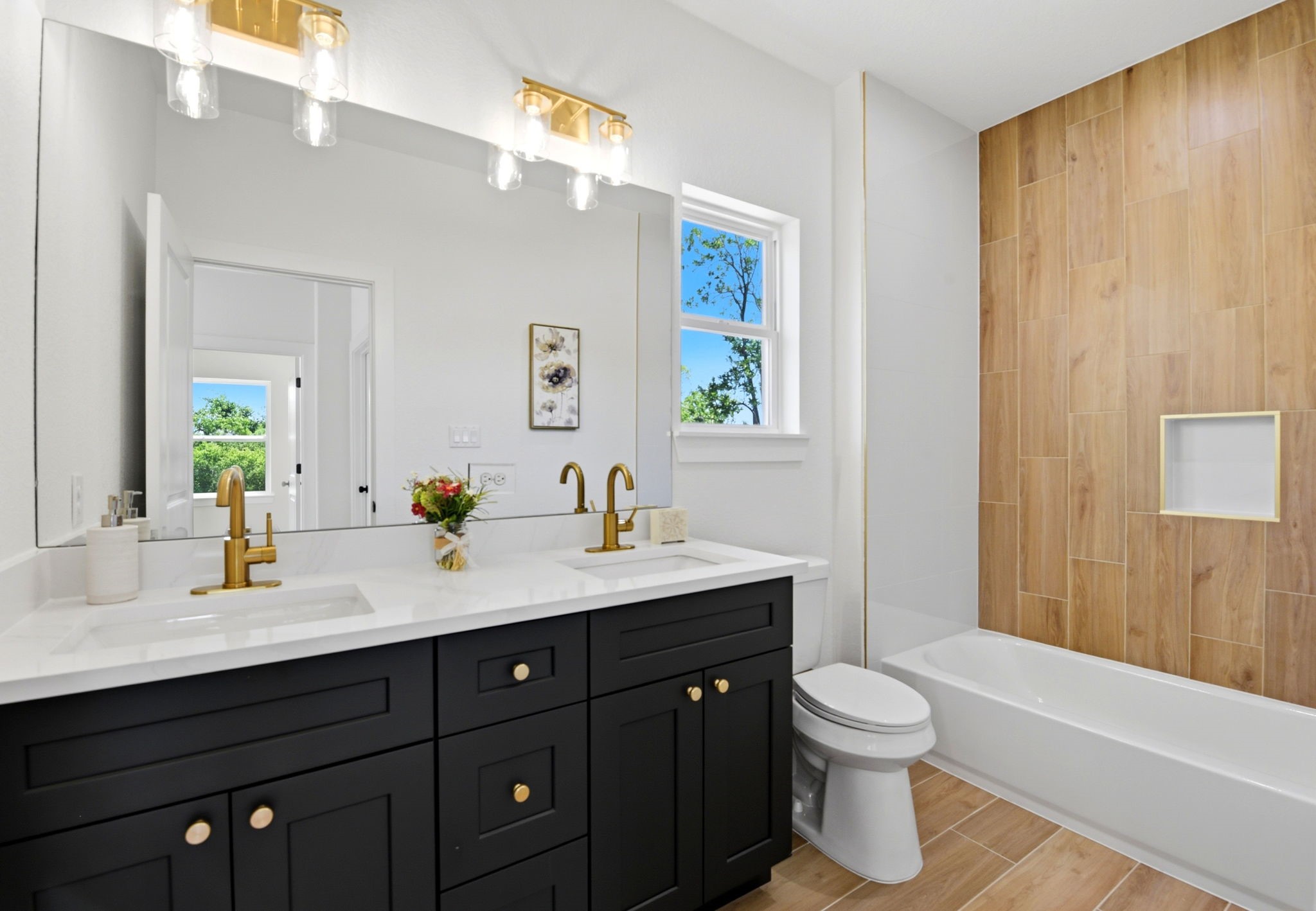 Modern secondary bathroom with dual sinks and sleek black cabinetry. Features gold fixtures, a large mirror, and a window for natural light. The bathtub area has wood-style tiles for a warm touch.