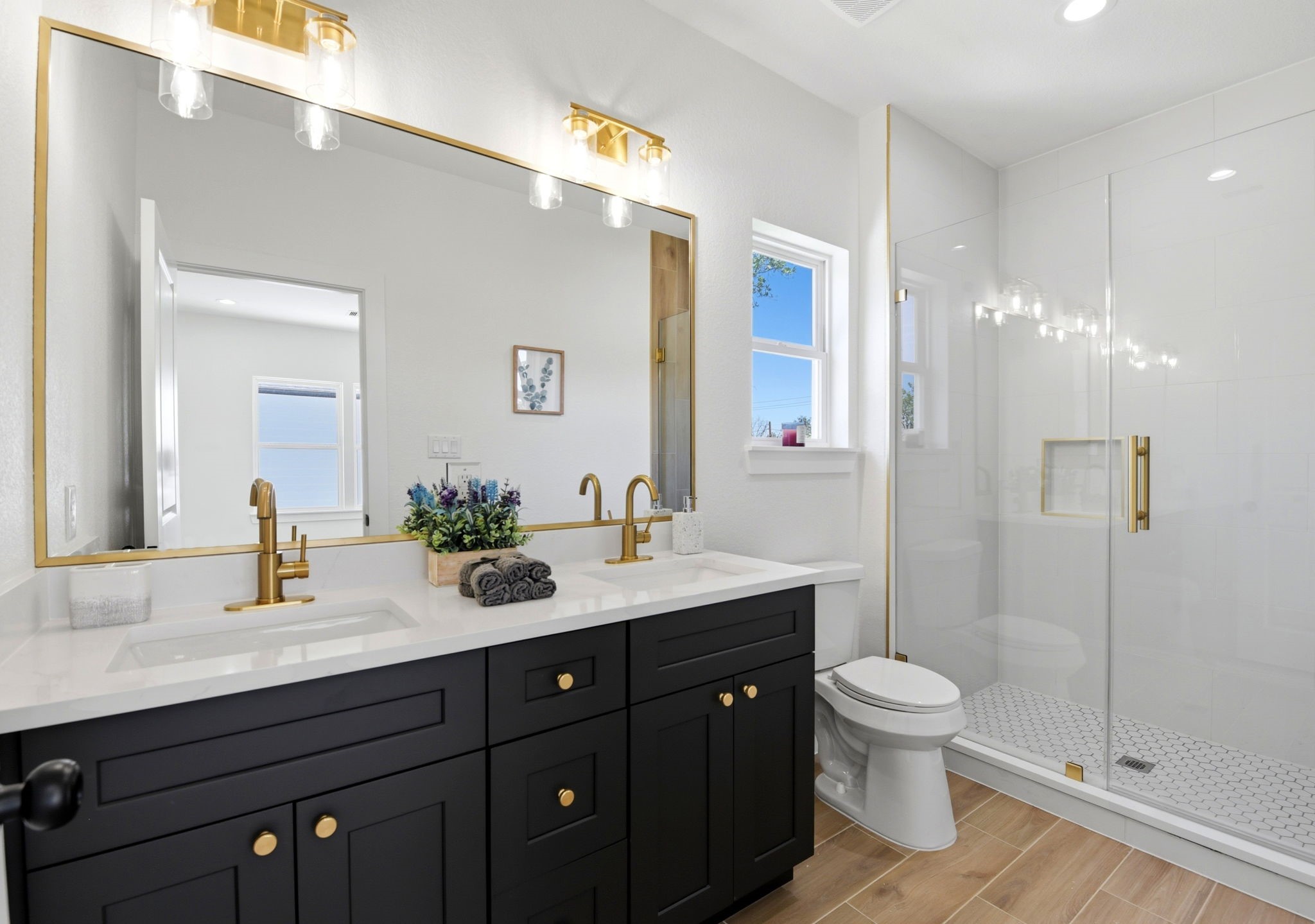 This modern primary bathroom features a spacious double vanity with sleek black cabinets and gold fixtures. It includes a large mirror, glass-enclosed shower, and bright lighting. The wood-like tile flooring and white walls add warmth and elegance.