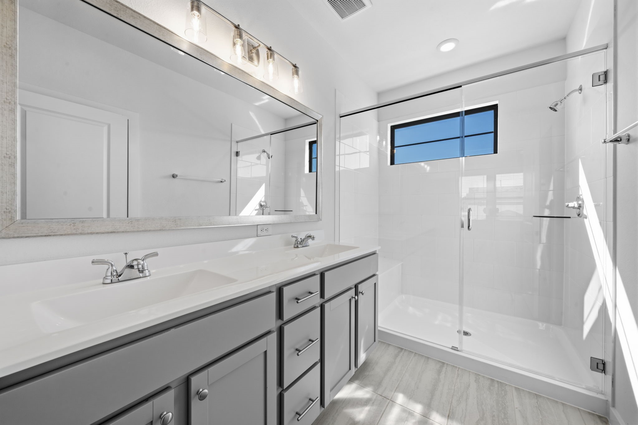 Welcome to a bright and beautifully finished primary bathroom, featuring a spacious walk-in glass shower, double vanity, sleek grey cabinetry, and modern finishes. Natural light pours in through the window, creating a fresh and inviting space to start and end your day.