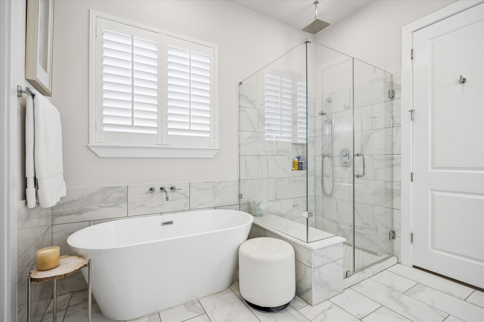 Enjoy a daily dose of luxury in this marble-wrapped primary bathroom with a freestanding soaking tub, oversized glass shower, wood shuttered window, and fresh modern finishes that feel calm and elevated.