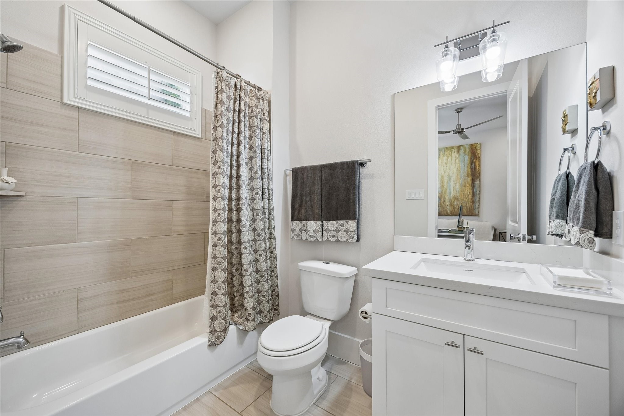 The guest ensuite bathroom is crisp and inviting with a clean white vanity, designer lighting, a tub and shower combo with tile surround, and a shuttered window that adds charm and natural light.