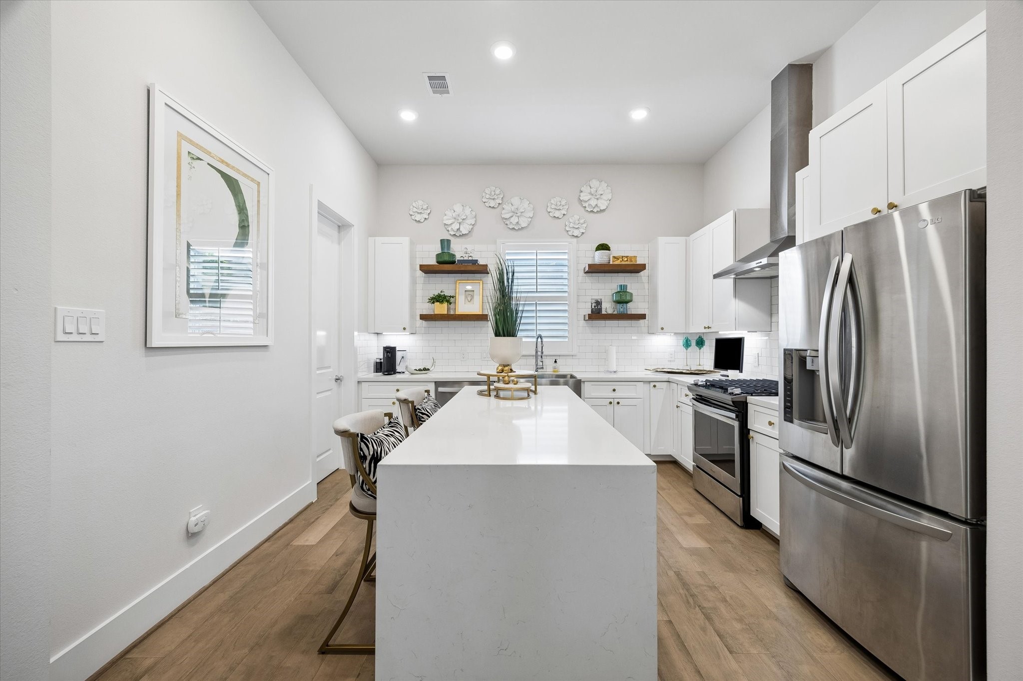 Crisp, clean, and ready to serve, this beautiful kitchen is filled with high-end style and everyday function, including a waterfall quartz island and countertops, subway tile backsplash, under-cabinet lights, stainless-steel appliances, a sleek hood vent, open shelving, modern cabinetry and a large walk-in pantry.