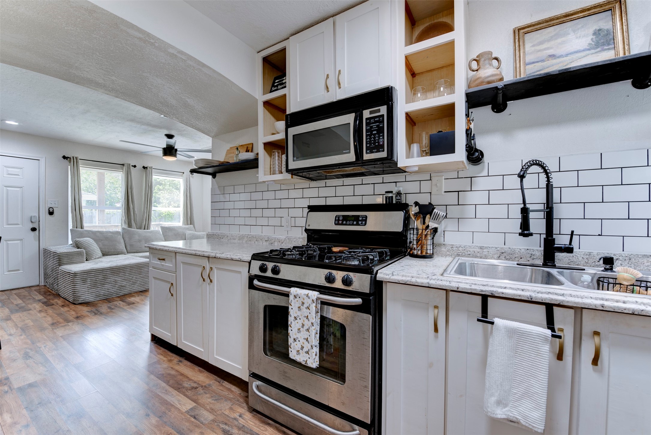 Kitchen to living room view showcasing GE gas range, stainless over-the-range microwave, matte black pull-down spring faucet, double-basin stainless sink, granite countertops, white subway tile backsplash with dark grout, white shaker cabinetry with brushed gold hardware, open pine cubbies, black pipe bracket floating shelf, dark wood-look LVP flooring, ceiling fan with light kit, and arched opening to living room.