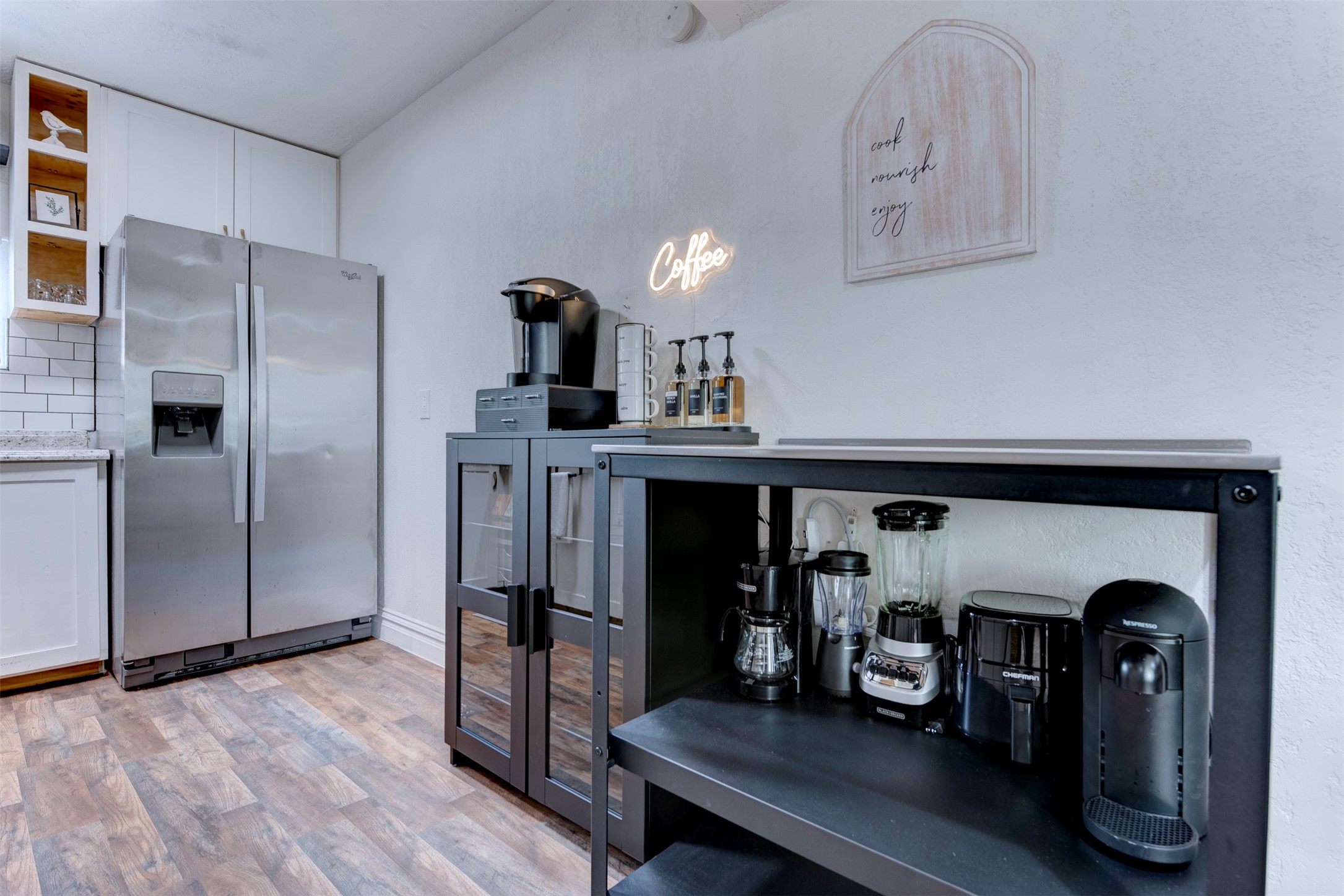Kitchen and coffee bar wide view showing Whirlpool stainless side-by-side refrigerator with ice and water dispenser, white shaker upper cabinets with natural pine open cubbies, white subway tile backsplash, granite countertops, and dedicated black metal coffee and appliance station with Keurig and Nespresso machines.