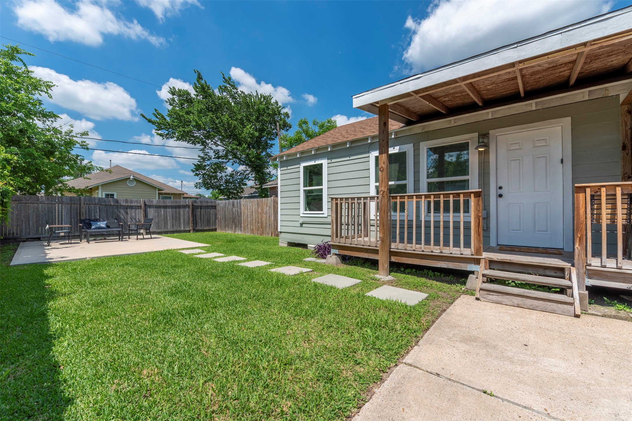 Exterior view featuring fresh sage-green lap siding, white trim, oversized single-pane windows, covered wood-porch with pendant light, concrete stepping stone path, and fully fenced yard.
