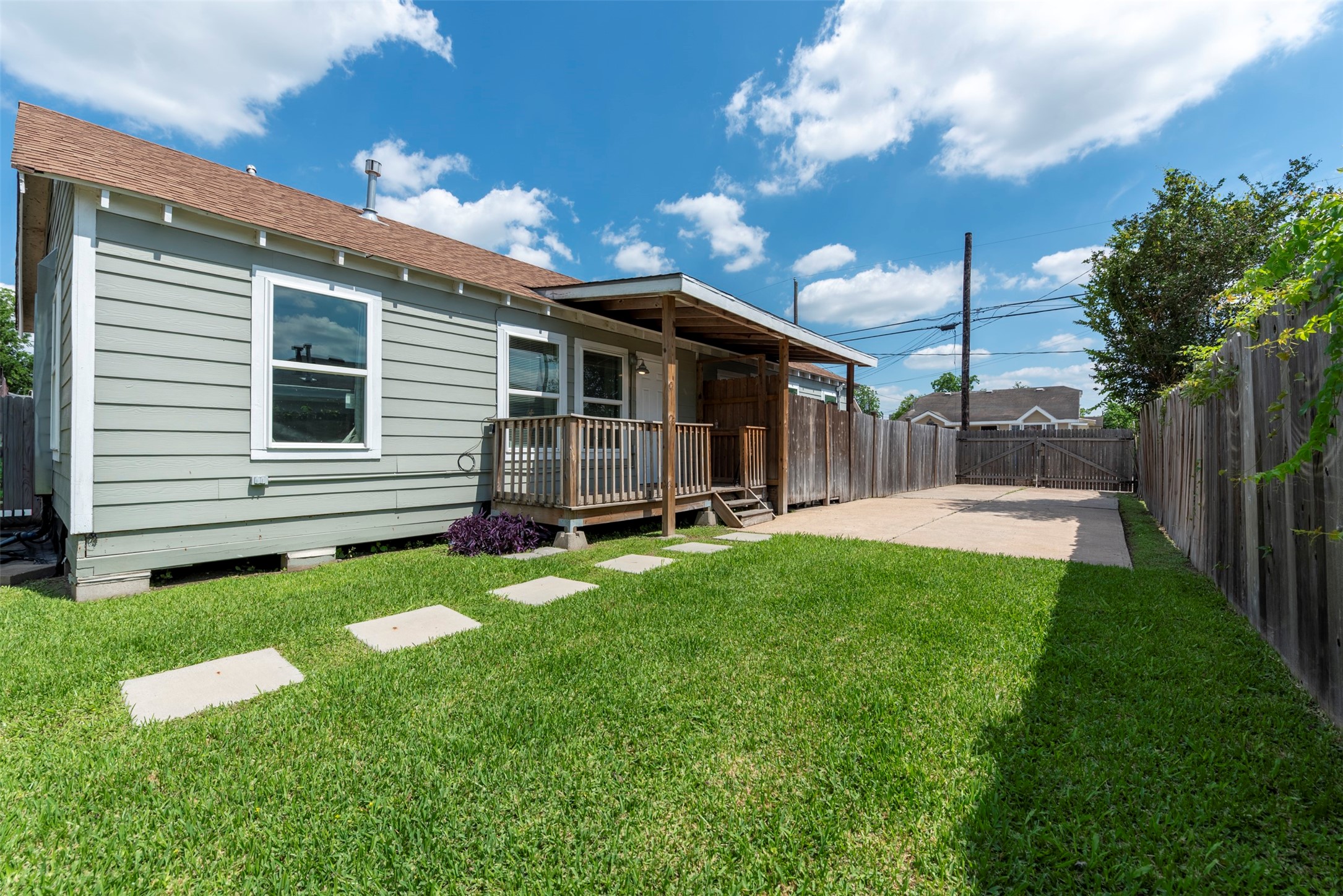 Side unit view showcasing sage-green lap siding, white-trimmed oversized windows, covered porch with wood post columns, concrete stepping stone path, and gated driveway access.