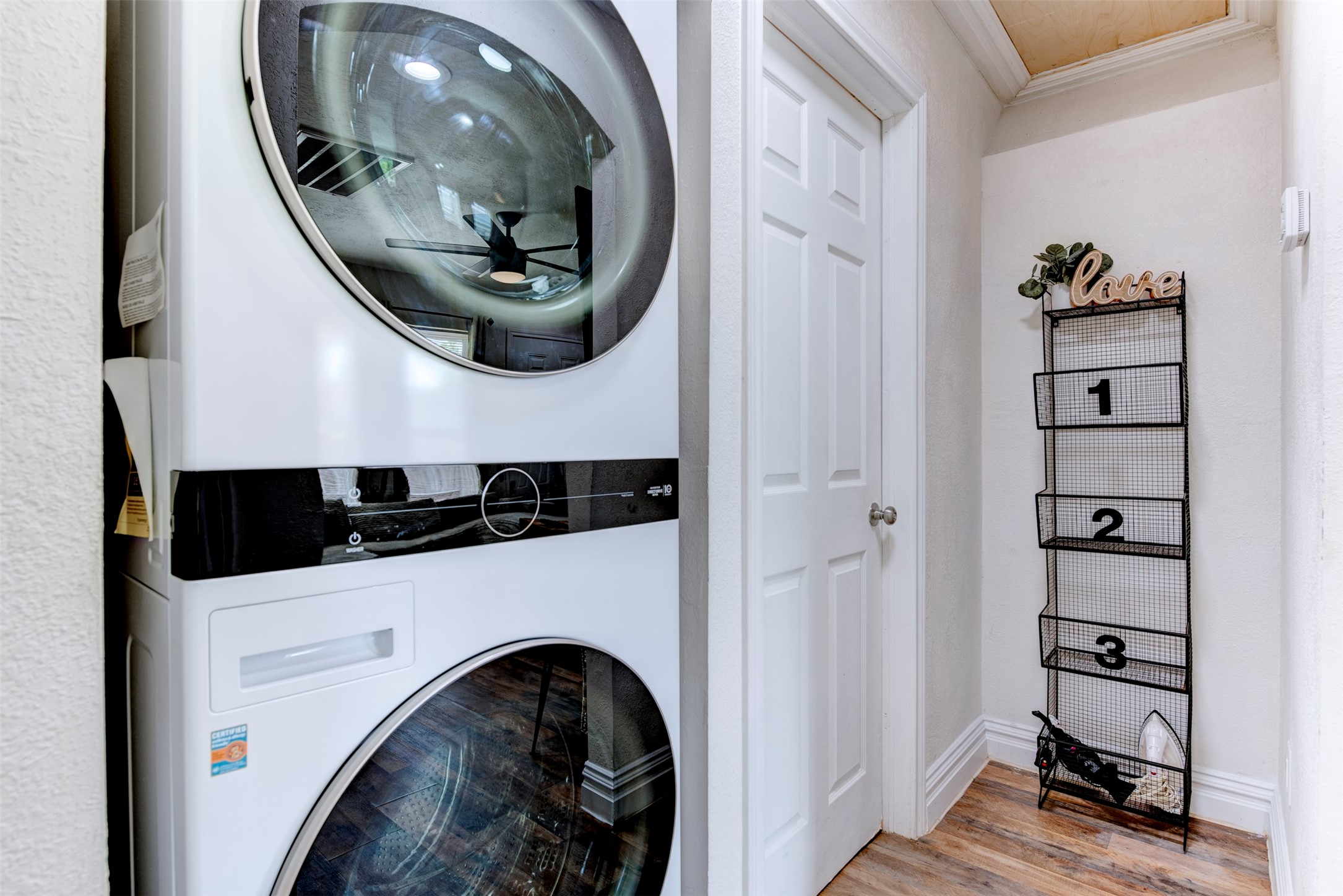 In-unit hallway laundry closet featuring full-size stacked front-load washer and dryer with black control panel accents, six-panel interior door, wood-look LVP flooring, and wood plank ceiling detail.