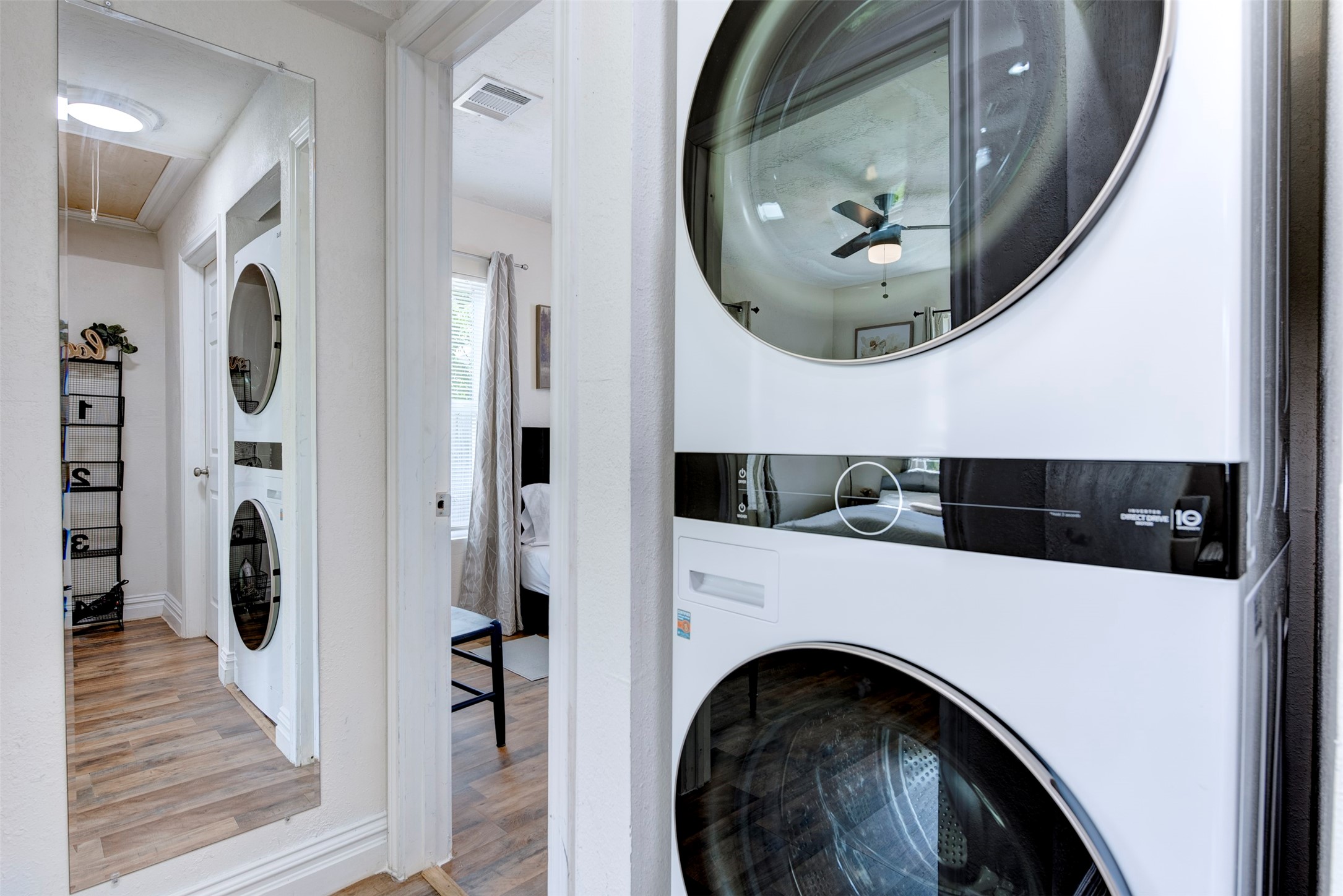 In-unit LG stacked front-load washer and dryer tucked into the hallway, with full-length mirror, wood-look flooring, and sightline directly into the bedroom.