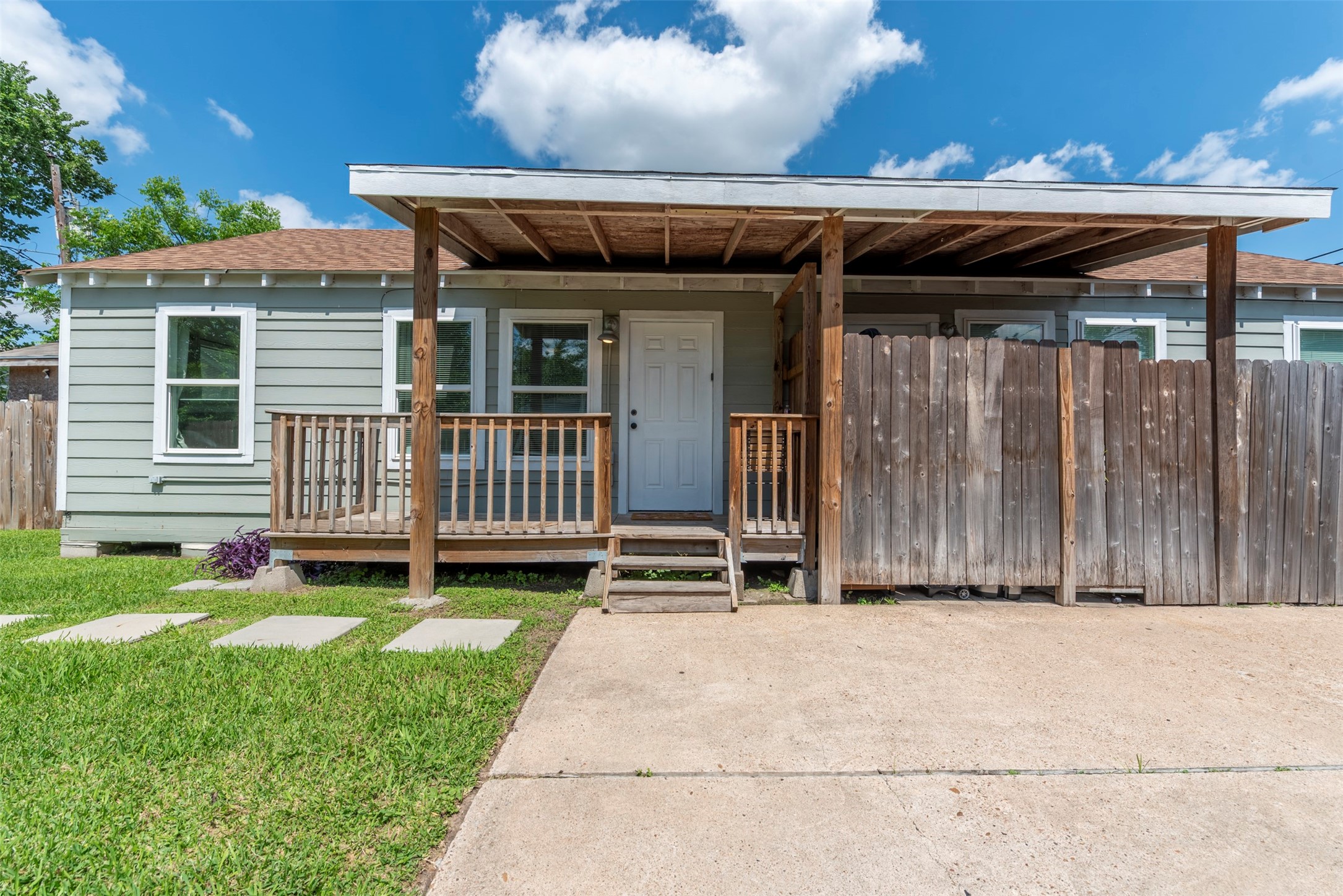 Front exterior with sage-green lap siding, white trim, covered front porch with exposed wood beam ceiling, wood railing, pendant wall light, and concrete driveway.