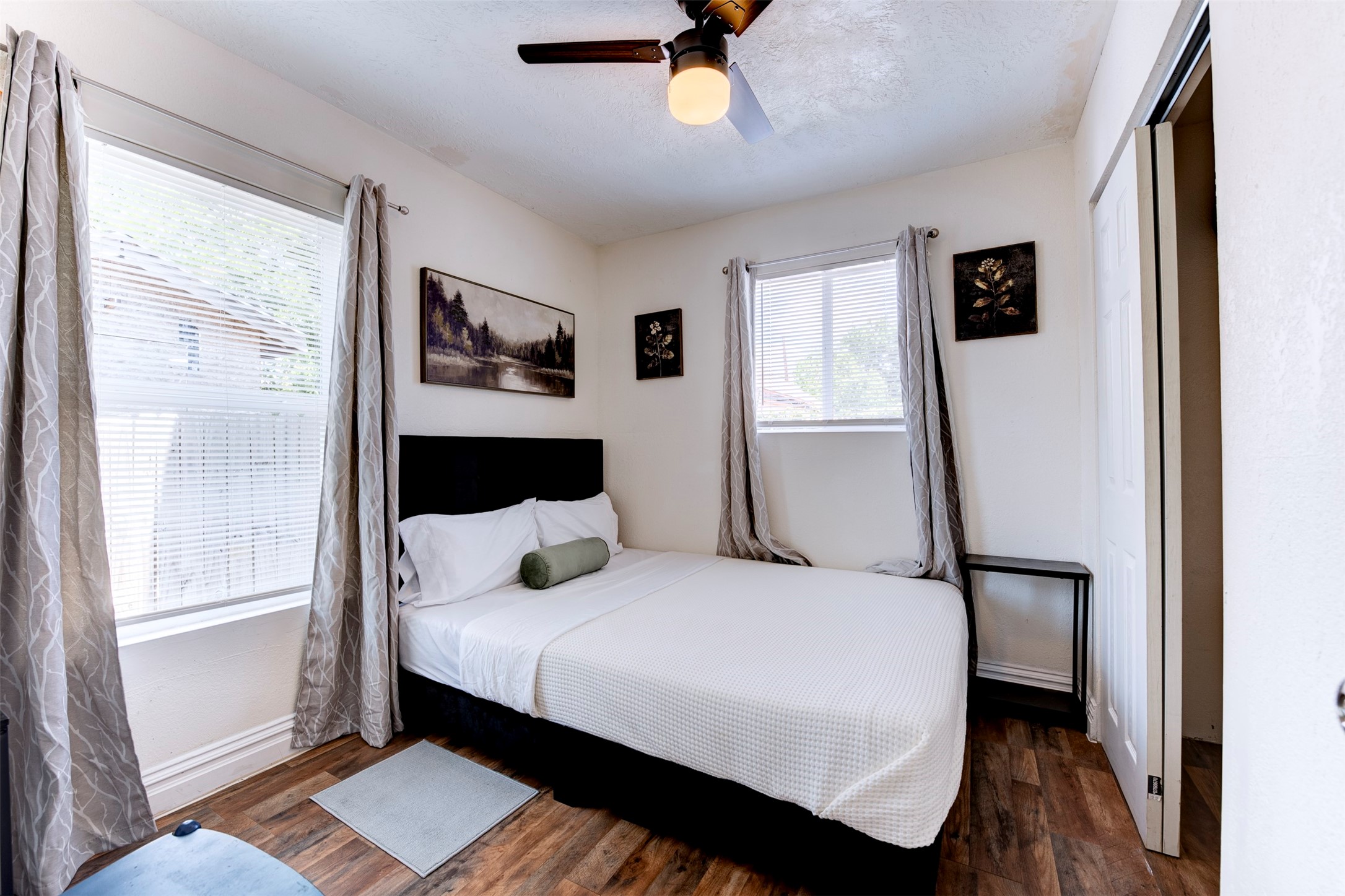 Bedroom featuring dark wood-look LVP flooring, ceiling fan with light kit, dual windows with blinds and grommet curtains, and a reach-in closet with sliding door.