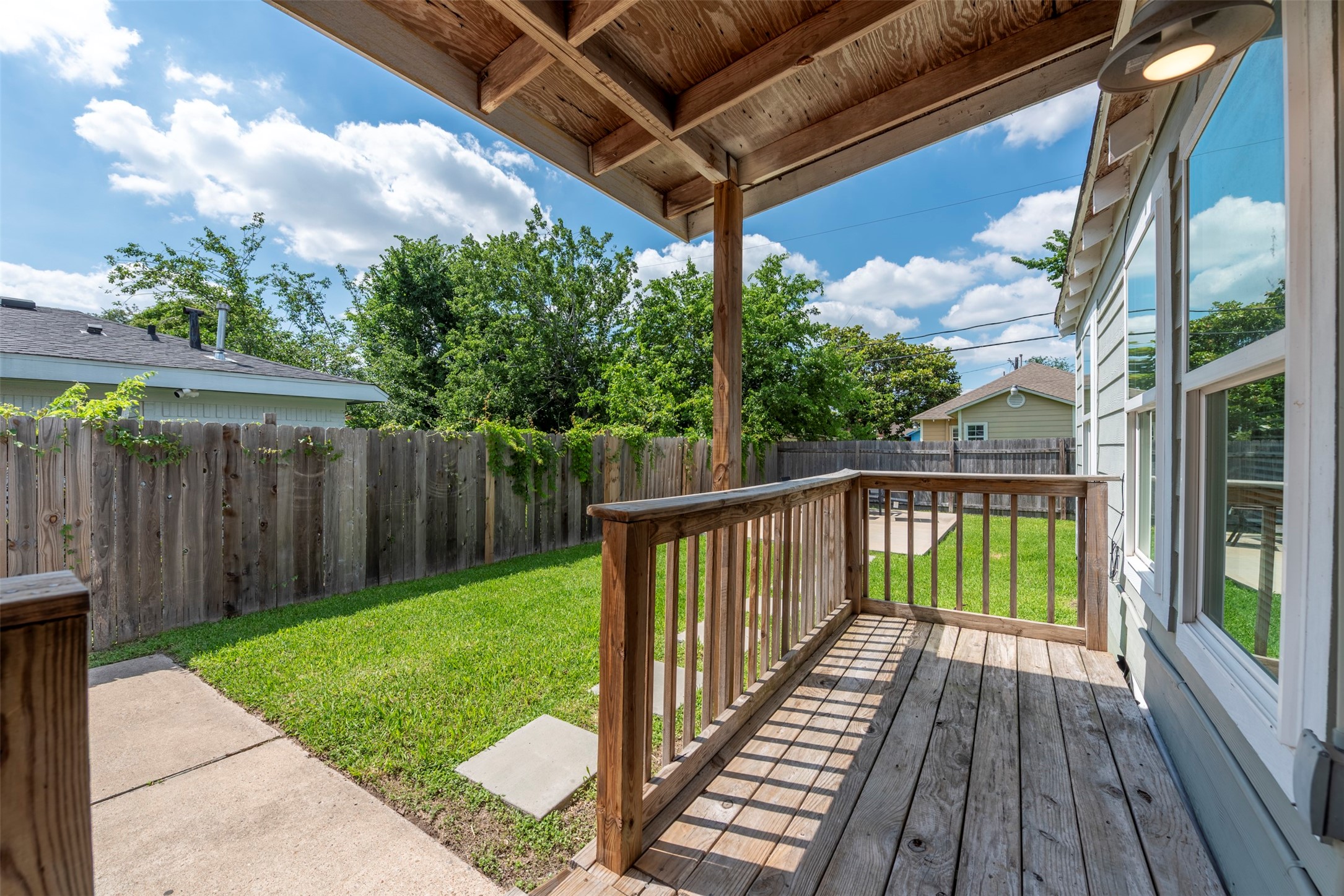 Covered front porch with exposed wood detail, pendant wall light, wood plank decking, wood baluster railing, and oversized single-pane window with direct sightlines to the fully fenced backyard.