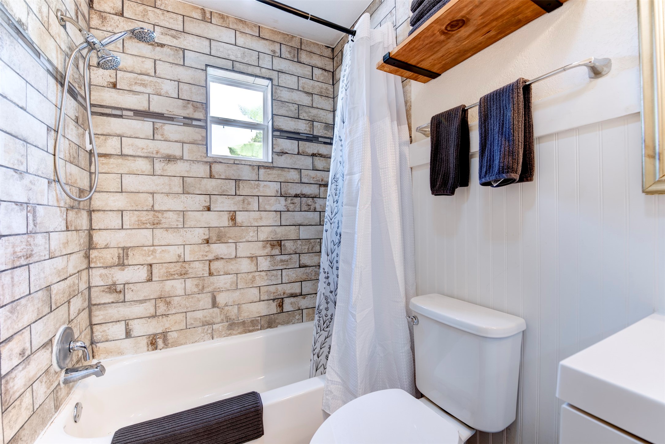 Tub and shower detail featuring floor-to-ceiling rustic brick-style large-format tile, chrome handheld shower head with slide bar, white soaking tub, operable window for natural light and ventilation, natural wood floating shelf with black iron rod, and beadboard wainscoting.