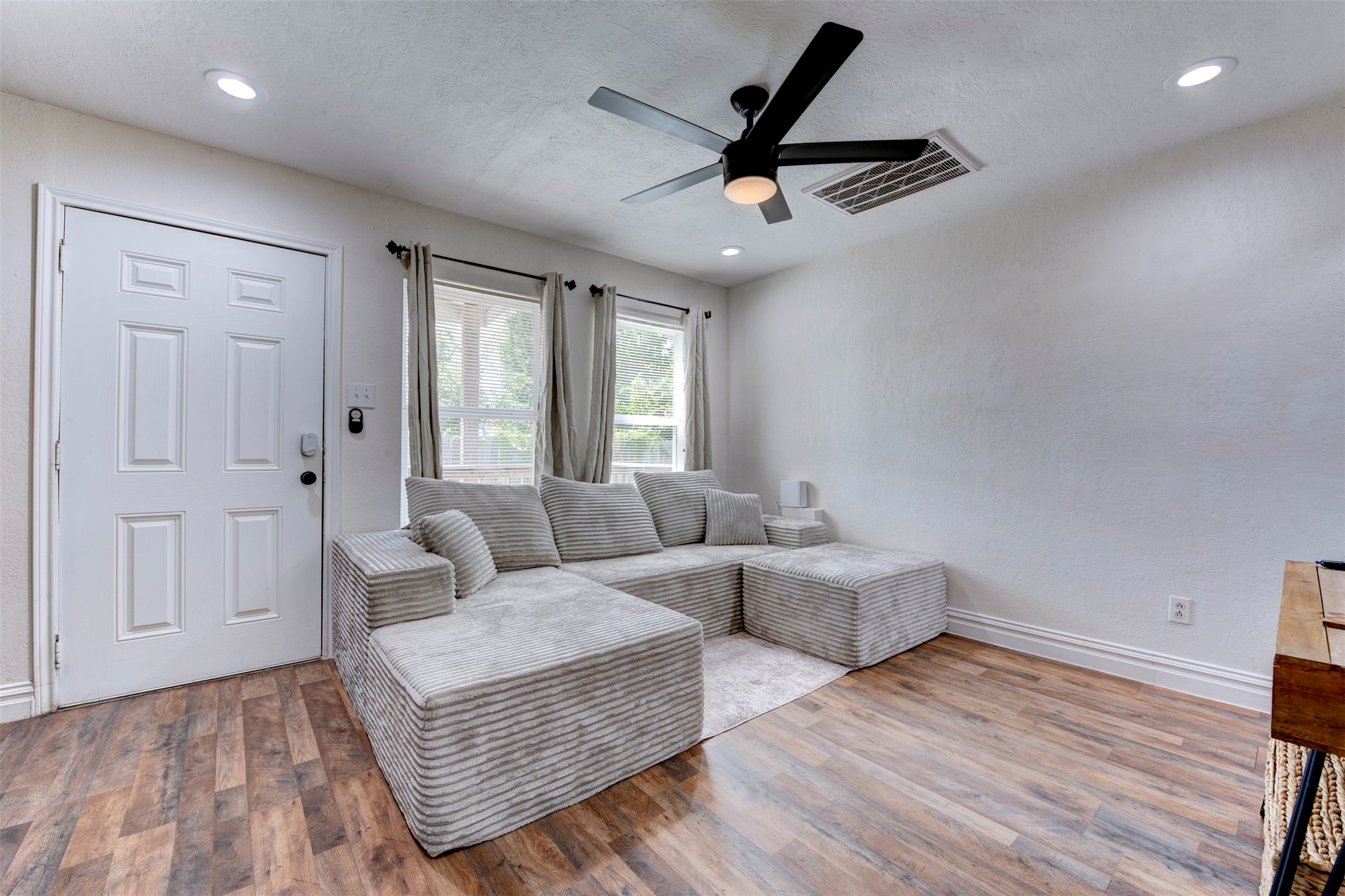 Living room wide angle showcasing white six-panel entry door with smart lock, oversized window with grommet linen curtains, wood-look LVP flooring, five-blade black ceiling fan with light kit, recessed lighting, and oversized returns with white baseboard trim throughout.