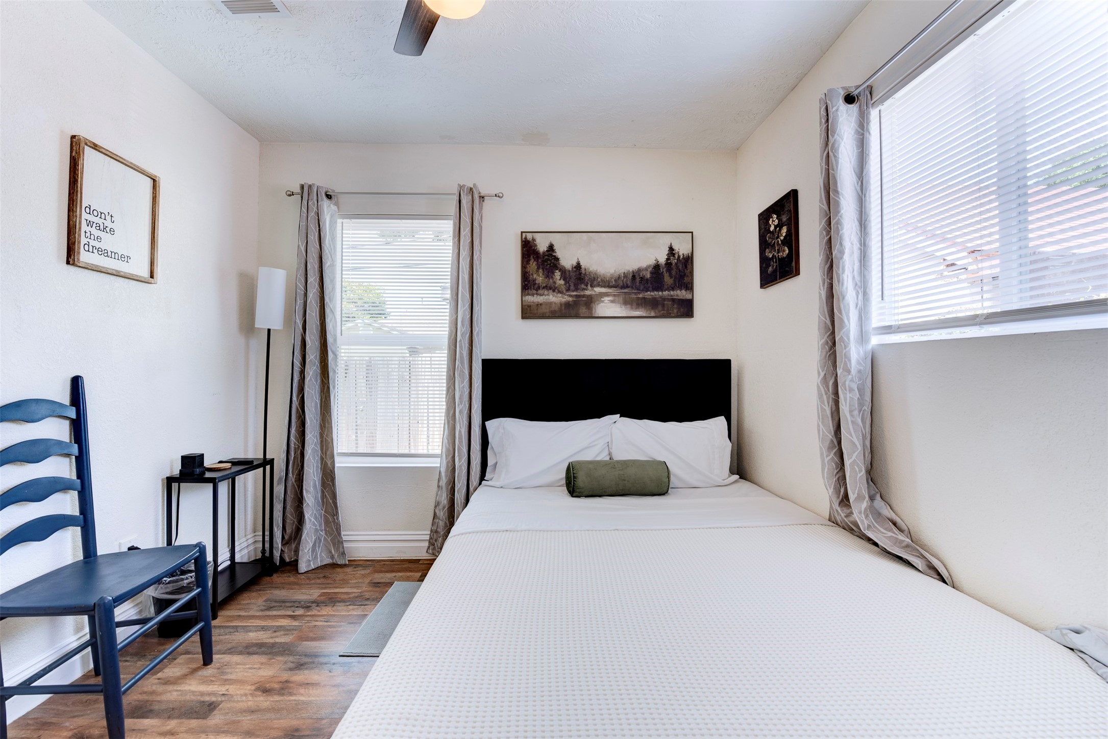 Alternate bedroom angle showing dual windows with natural light, dark wood-look LVP flooring, ceiling fan, and grommet curtain panels on black rod hardware.