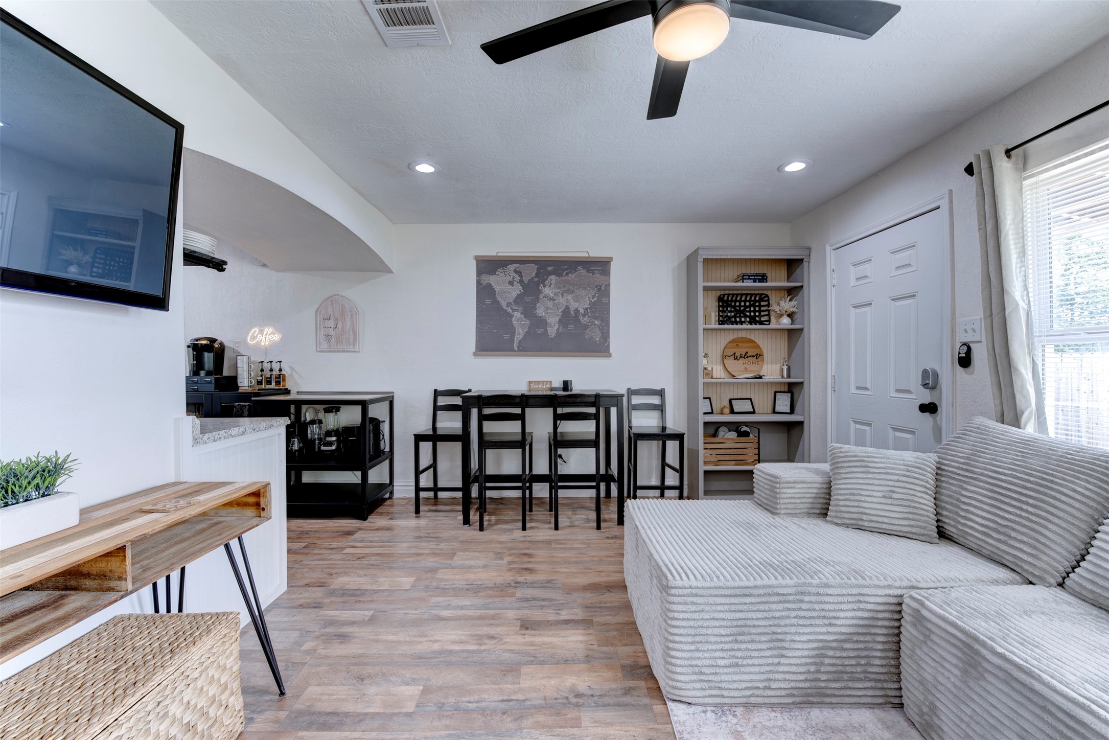 Full living and dining layout showing wood-look LVP flooring, five-blade black ceiling fan with light kit, recessed lighting, wall-mounted flat screen TV, built-in five-shelf bookcase with beadboard back panel, black bar-height dining set, dedicated coffee station with neon sign, white six-panel entry door with smart lock, and oversized window with natural light.