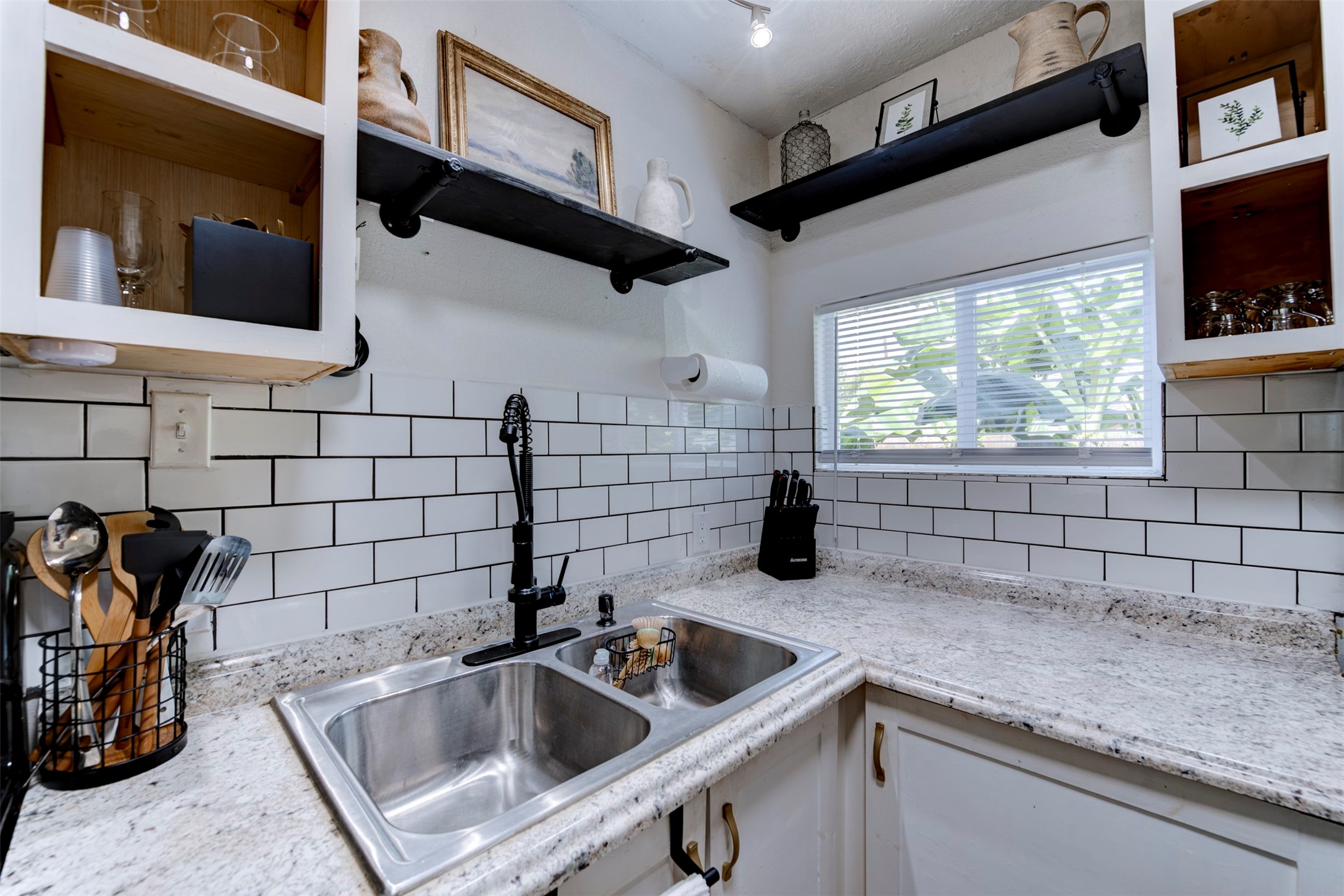 Kitchen sink area featuring double-basin stainless steel undermount sink, matte black pull-down spring faucet, granite countertops, white subway tile backsplash with dark grout, track lighting, open wood cabinets with natural pine interiors, and dual black pipe bracket floating shelves.