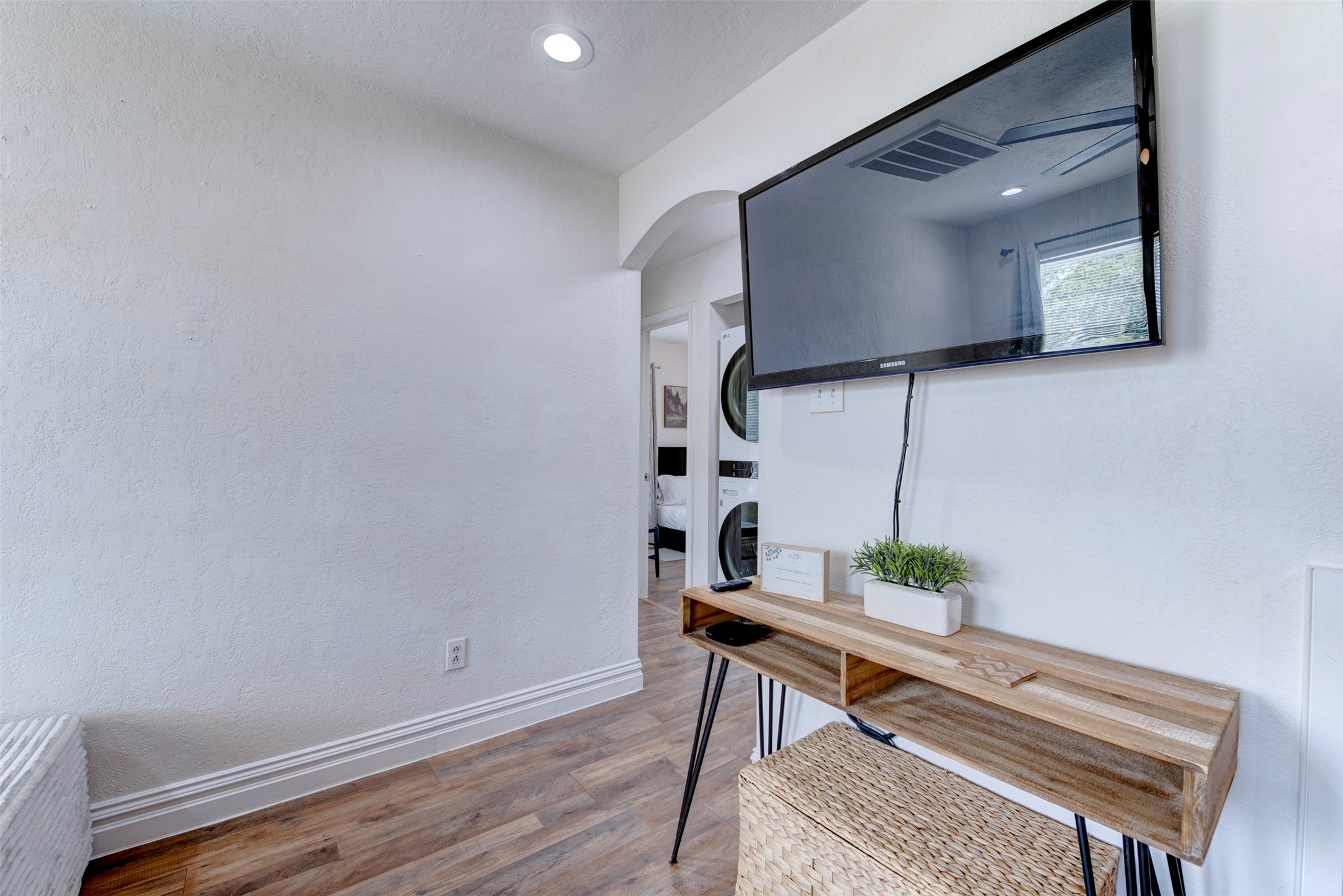 Living room TV wall detail featuring wall-mounted Samsung flat screen TV, natural wood hairpin leg console table, recessed lighting, arched hallway leading to bedrooms and in-unit stacked laundry, and wood-look LVP flooring throughout.
