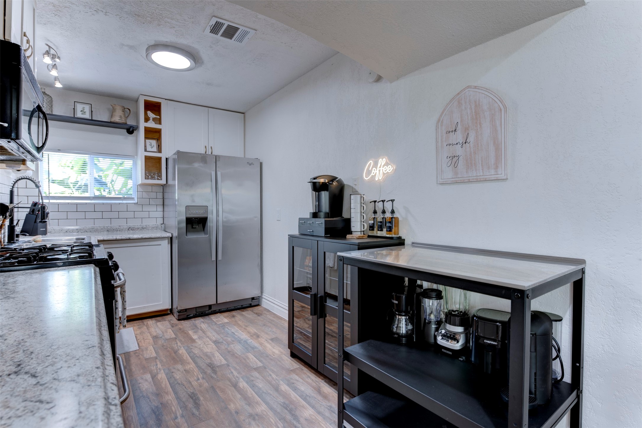 Kitchen wide view with GE gas range, granite countertops, Whirlpool stainless side-by-side refrigerator with ice and water dispenser, white subway tile backsplash, white shaker upper cabinetry, open pine cubbies, matte black pull-down faucet, track lighting, flush mount ceiling light, and dedicated black metal coffee and appliance station with Keurig brewer and syrup dispensers.
