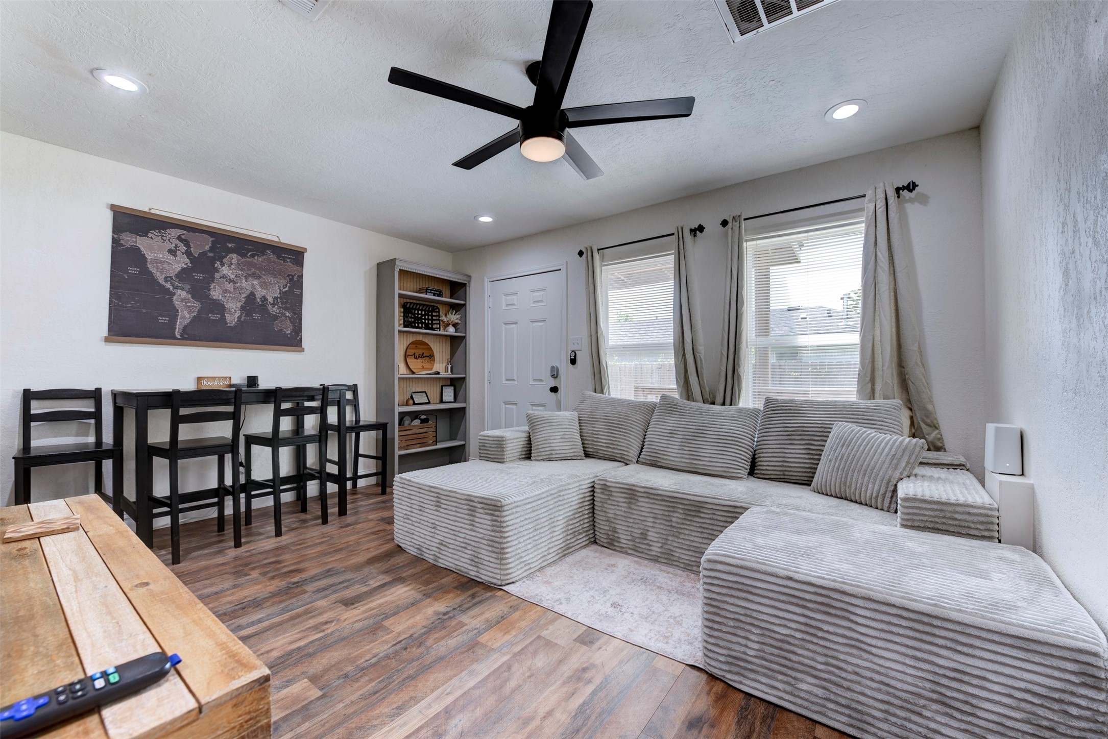 Living and dining area featuring dark wood-look LVP flooring, oversized black ceiling fan with light kit, recessed lighting, dual oversized windows with grommet linen curtain panels on black rod hardware, five-shelf built-in bookcase with beadboard back panel, six-panel entry door with smart lock, and black bar-height dining set.