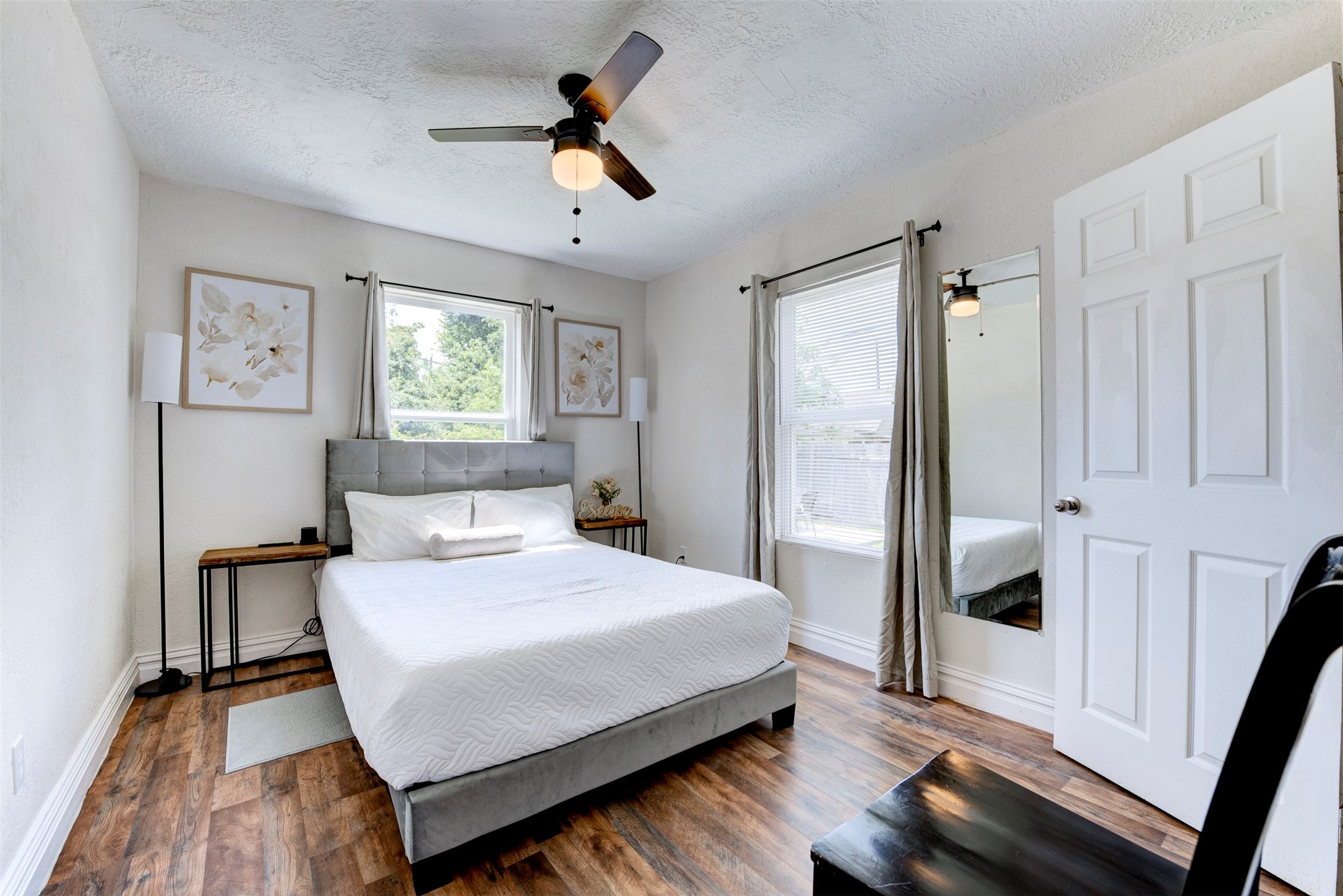 Primary bedroom with dark wood-look LVP flooring, ceiling fan with light kit, two windows with grommet curtain panels, full-length mirror, and door leading to private bath.