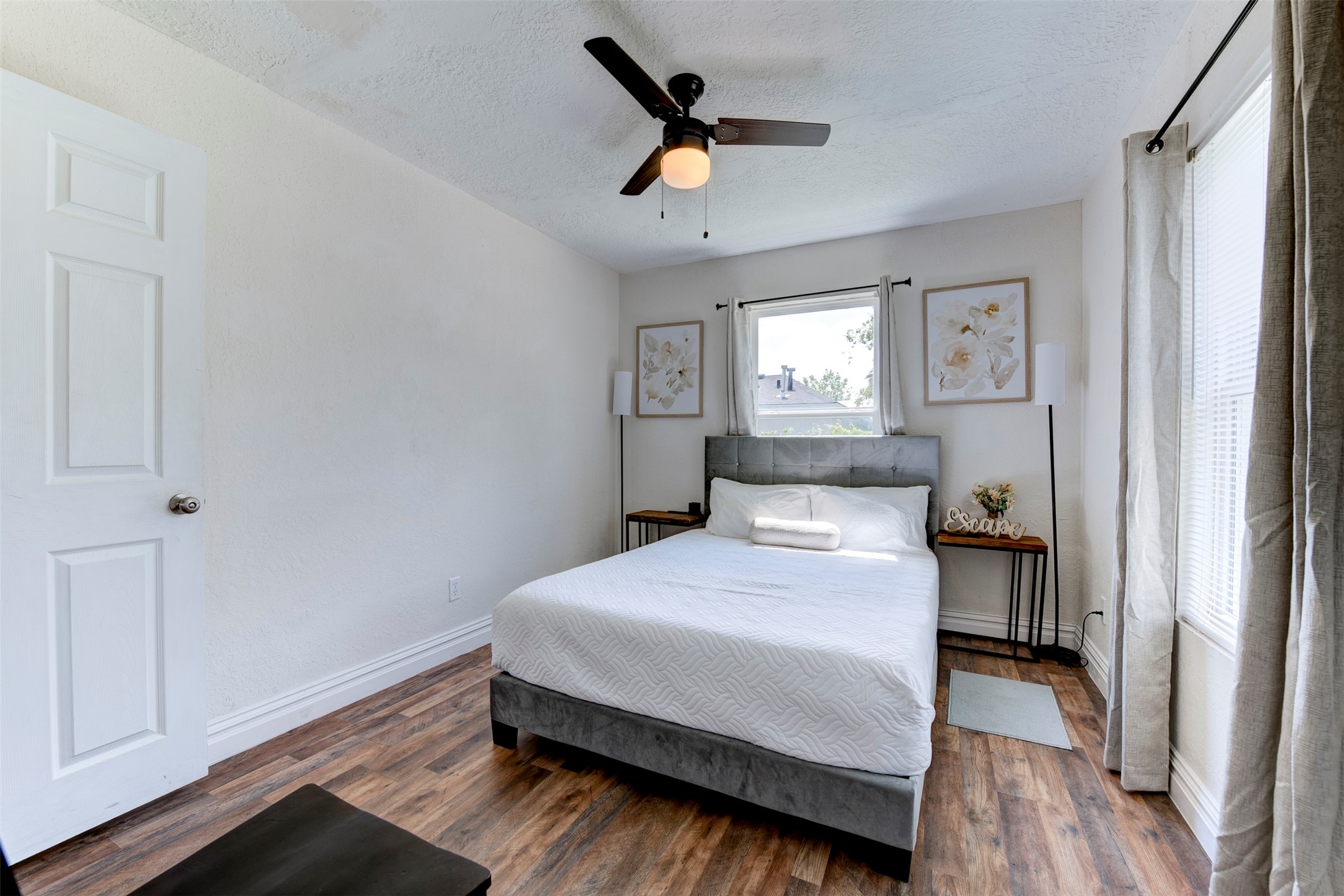 Second bedroom with dark wood-look LVP flooring, ceiling fan with light kit, oversized window with grommet curtains on black rod, and six-panel interior door.