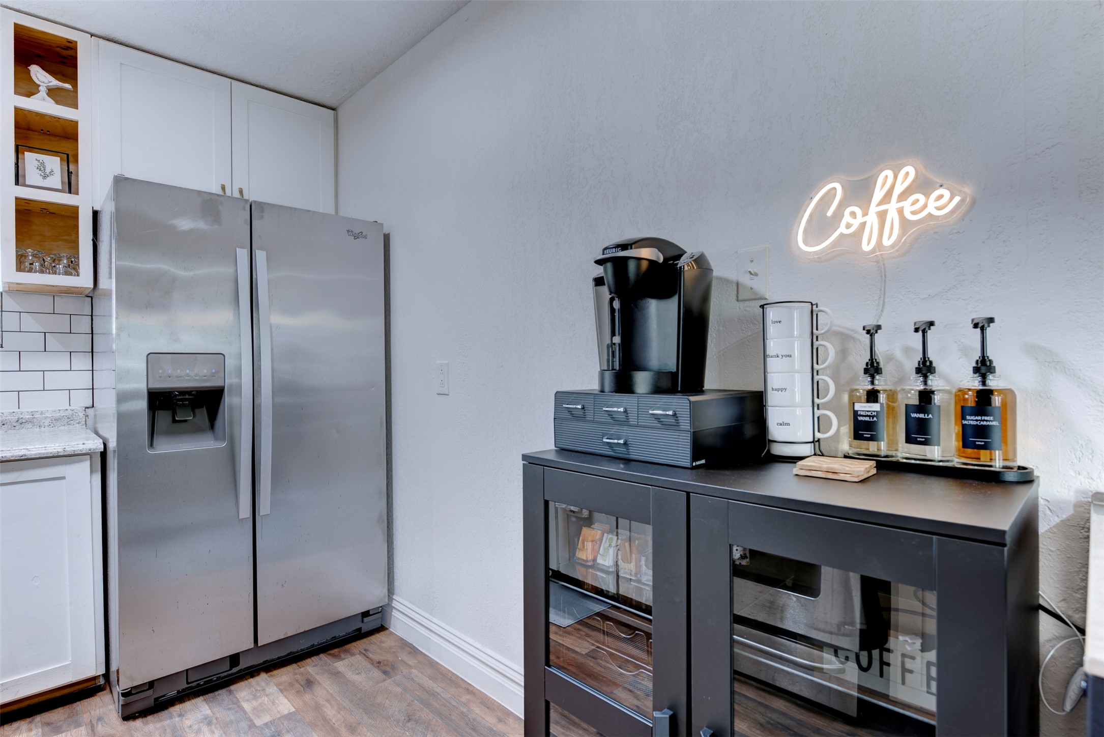 Coffee and appliance station with Keurig brewer, labeled syrup dispensers, white shaker cabinetry, and open pine cubby shelving