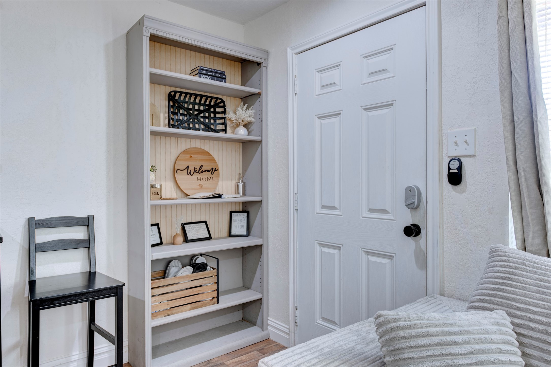 Entryway featuring white six-panel door with smart lock and black lever hardware, built-in five-shelf bookcase with natural pine beadboard back panel, wood-look LVP flooring, and oversized window with natural light.