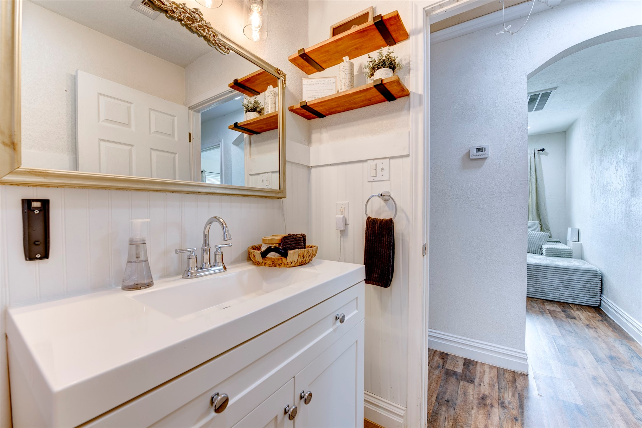 Bathroom vanity close-up showing white shaker cabinetry with chrome knobs, single-basin white countertop, chrome bridge faucet, ornate silver-framed mirror, dual natural wood floating shelves with black iron brackets, beadboard wainscoting, and open arched hallway leading to bedroom.