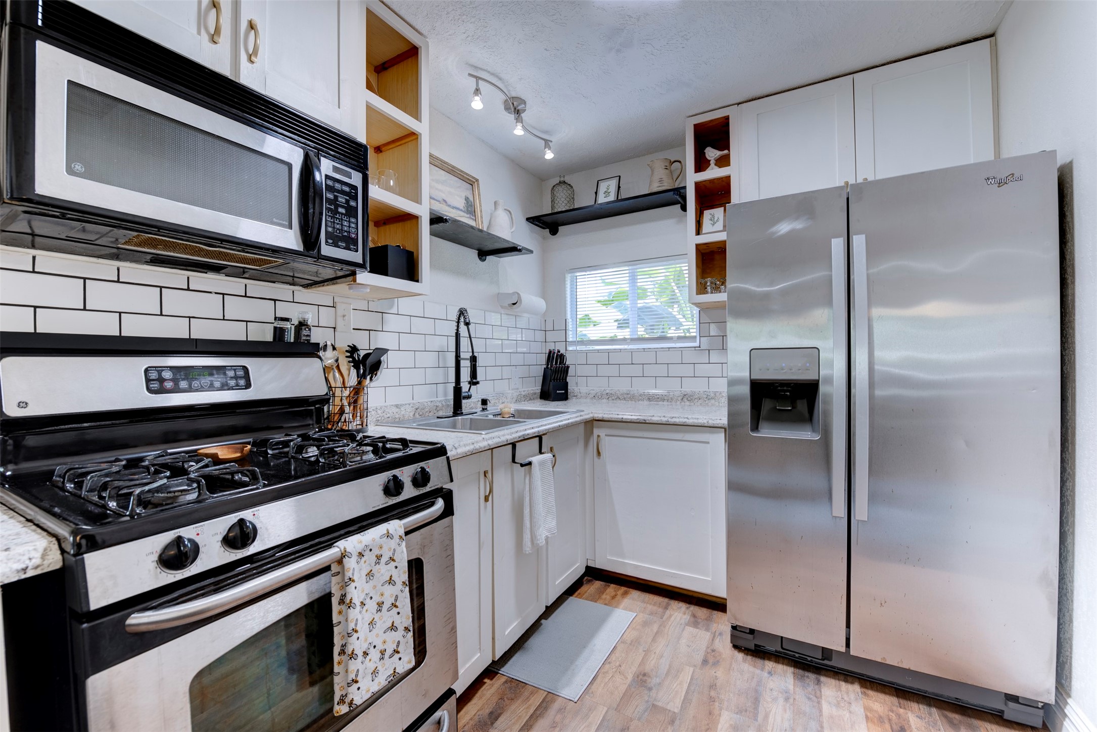 Full kitchen view featuring GE five-burner gas range with stainless finish, over-the-range stainless microwave, Whirlpool side-by-side refrigerator with ice and water dispenser, white shaker cabinetry with brushed gold hardware, granite countertops, white subway tile backsplash with dark grout, open pine cubby shelving, black pipe bracket floating shelf, track lighting, and operable window.