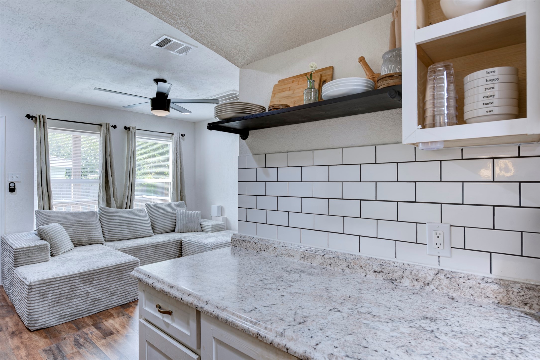 Kitchen pass-through view showing granite countertops with white and gray veining, white subway tile backsplash with dark grout, black steel open shelving with pipe brackets, white shaker cabinetry with brushed gold hardware, ceiling fan with light kit, and oversized window with grommet curtain panels.