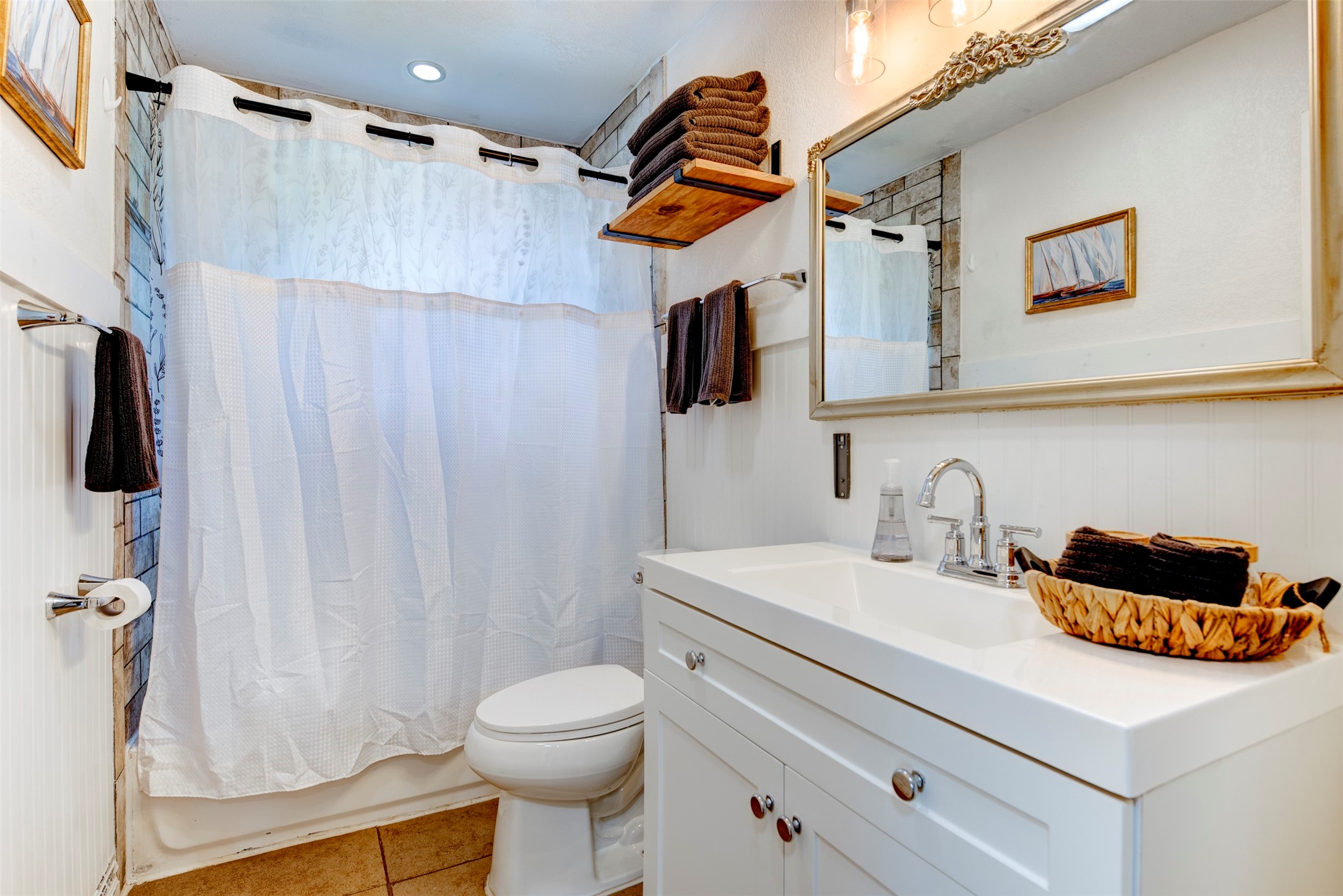 Full bathroom featuring white shaker vanity with chrome two-handle faucet, ornate silver-framed mirror, Edison bulb pendant lighting, open natural wood floating shelf, beadboard wainscoting, recessed ceiling light, and tiled shower surround.