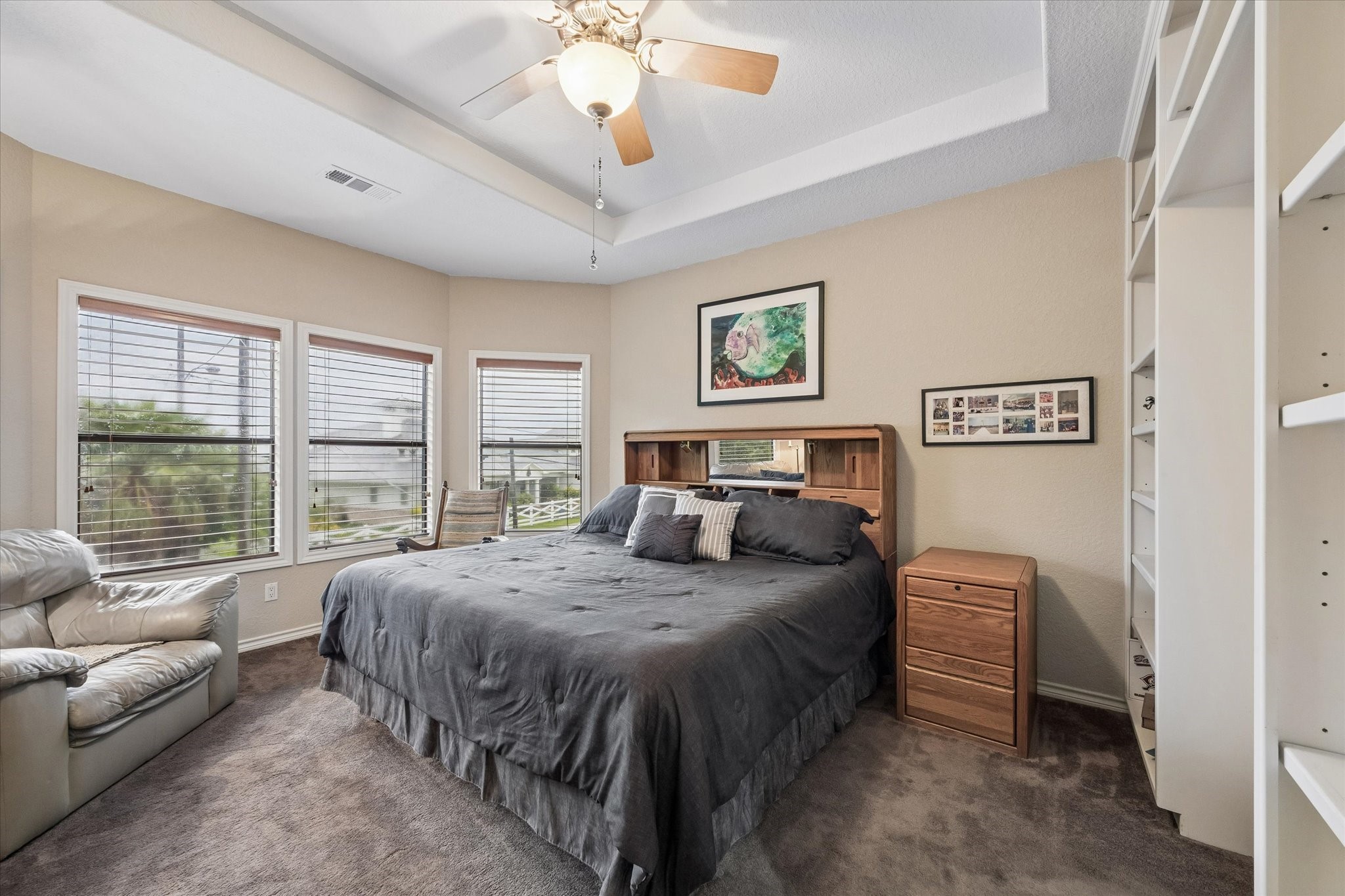 Spacious primary bedroom with tray ceiling, ceiling fan, and abundant natural light from multiple windows. Generous layout accommodates large furnishings with comfortable seating area.