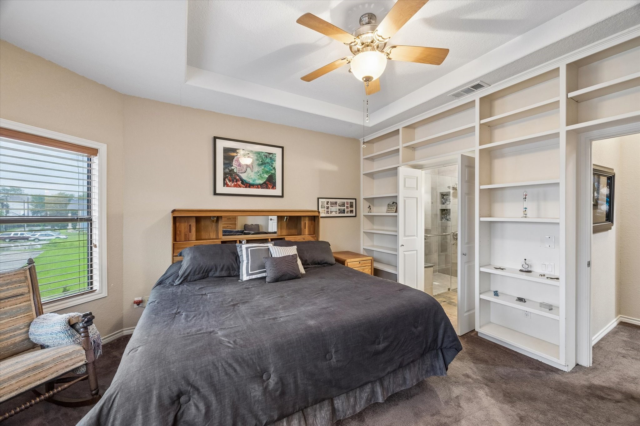 Primary bedroom featuring tray ceiling, ceiling fan, and custom built-in shelving. Direct access to the en-suite bath enchances functionaility and storage.