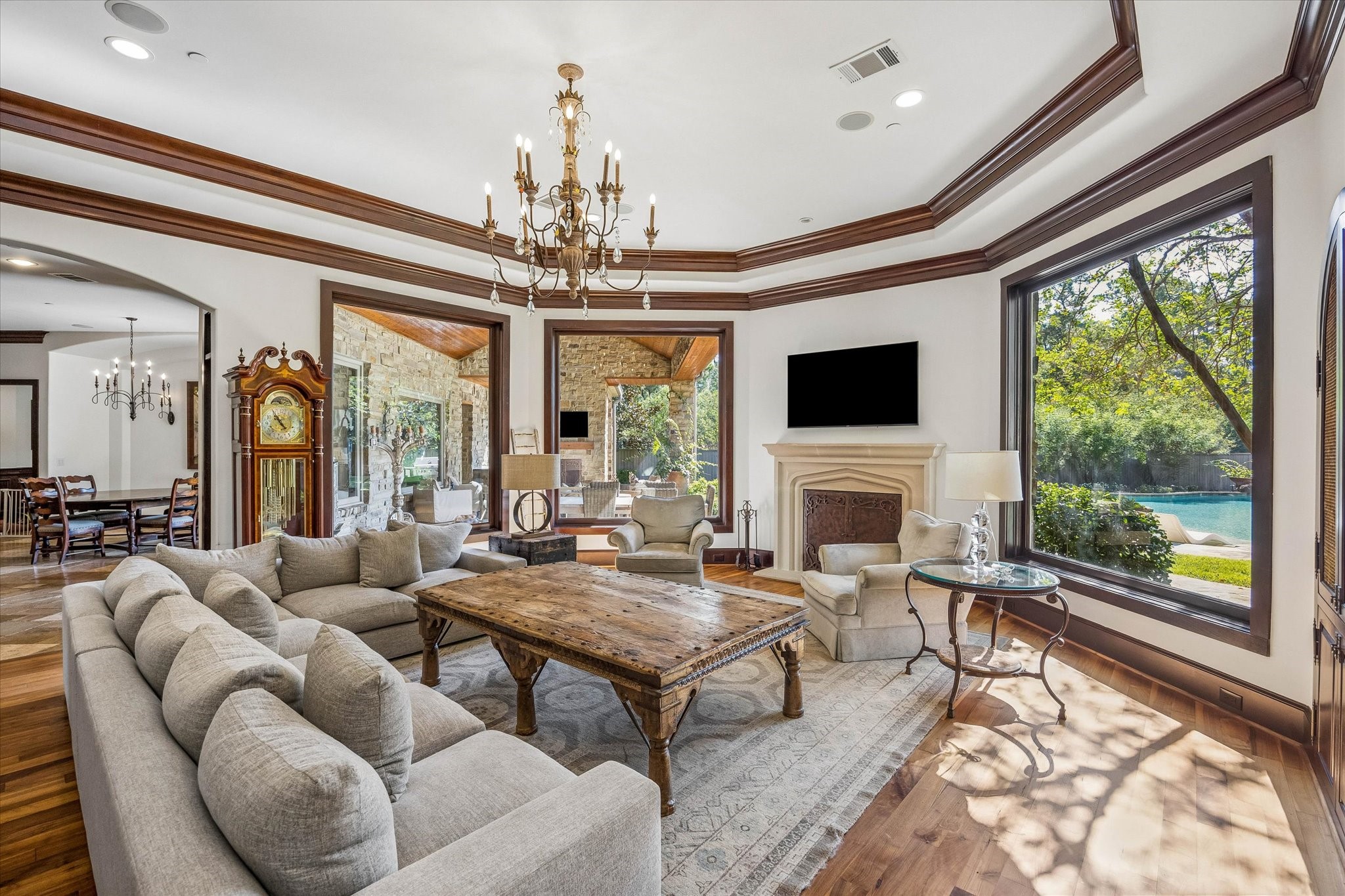 The family room is filled with natural light from expansive windows overlooking the pool and covered patio, while a fireplace, hardwood floors, and rich wood trim add warmth and character.