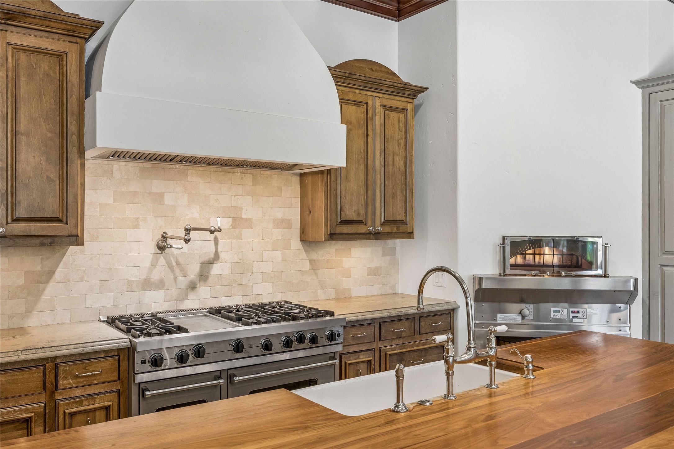 A chefs kitchen featuring a Viking gas range, custom vent hood, pot filler, and stone backsplash, complemented by a generous wood-topped island with farmhouse sink.