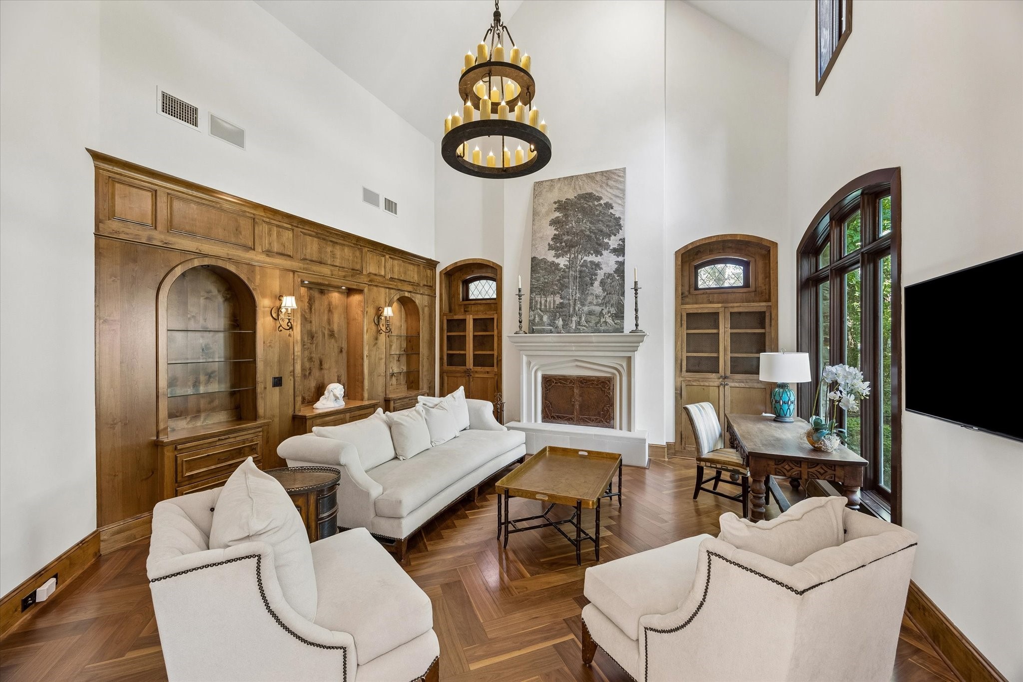 Impressive living room featuring soaring ceilings, rich wood-paneled walls with arched built-ins, a carved fireplace mantel, herringbone wood floors, and tall arched windows providing abundant natural light.